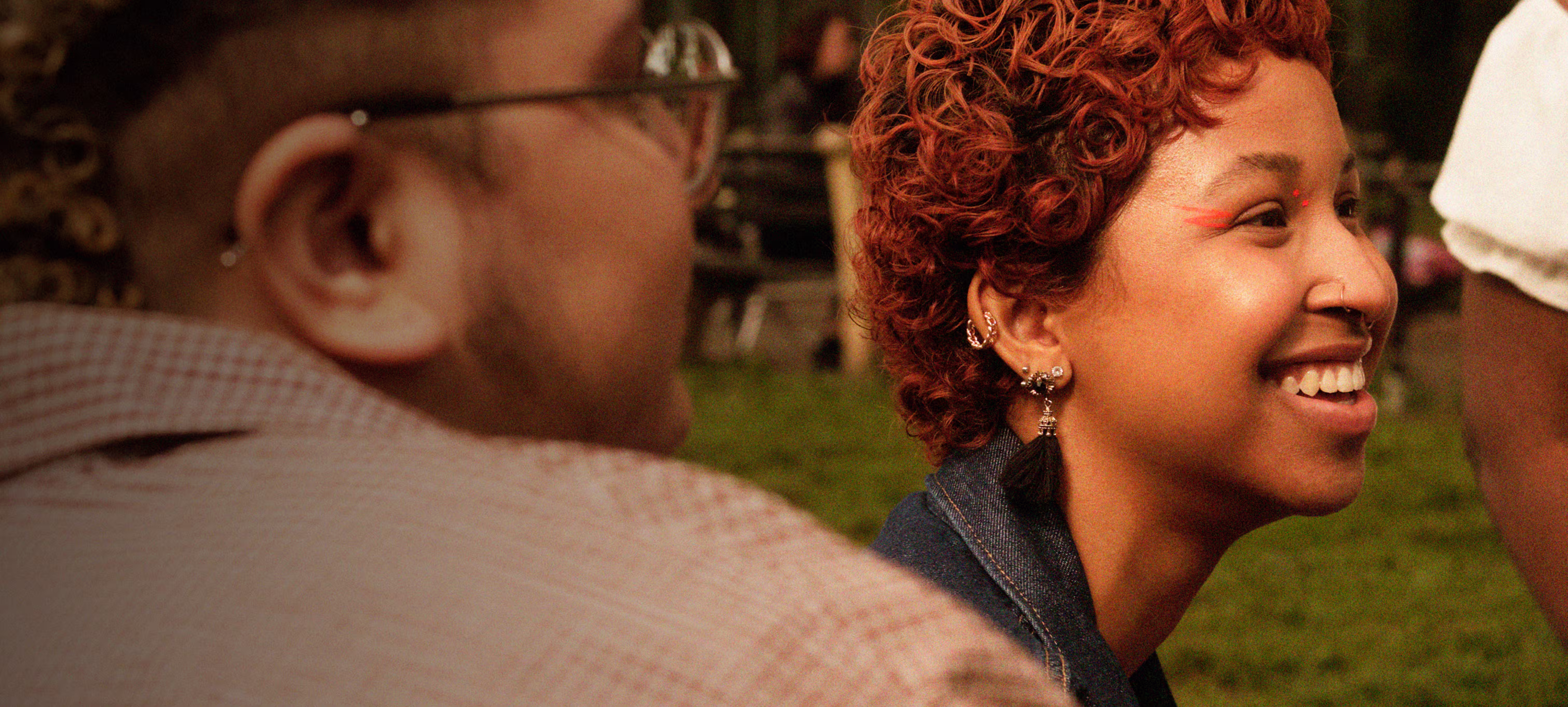 Side profile of a smiling person with curly red hair, face paint, and multiple earrings, sitting outdoors with another person nearby.