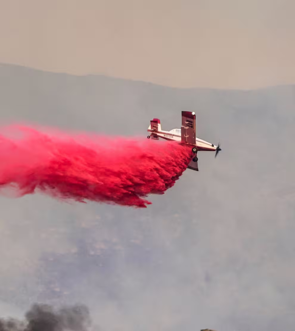 Small aircraft releasing a large plume of red smoke during flight over a mountainous area.