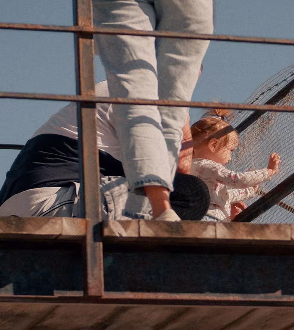 A toddler playing with a netted fence on a wooden platform while adults stand nearby.