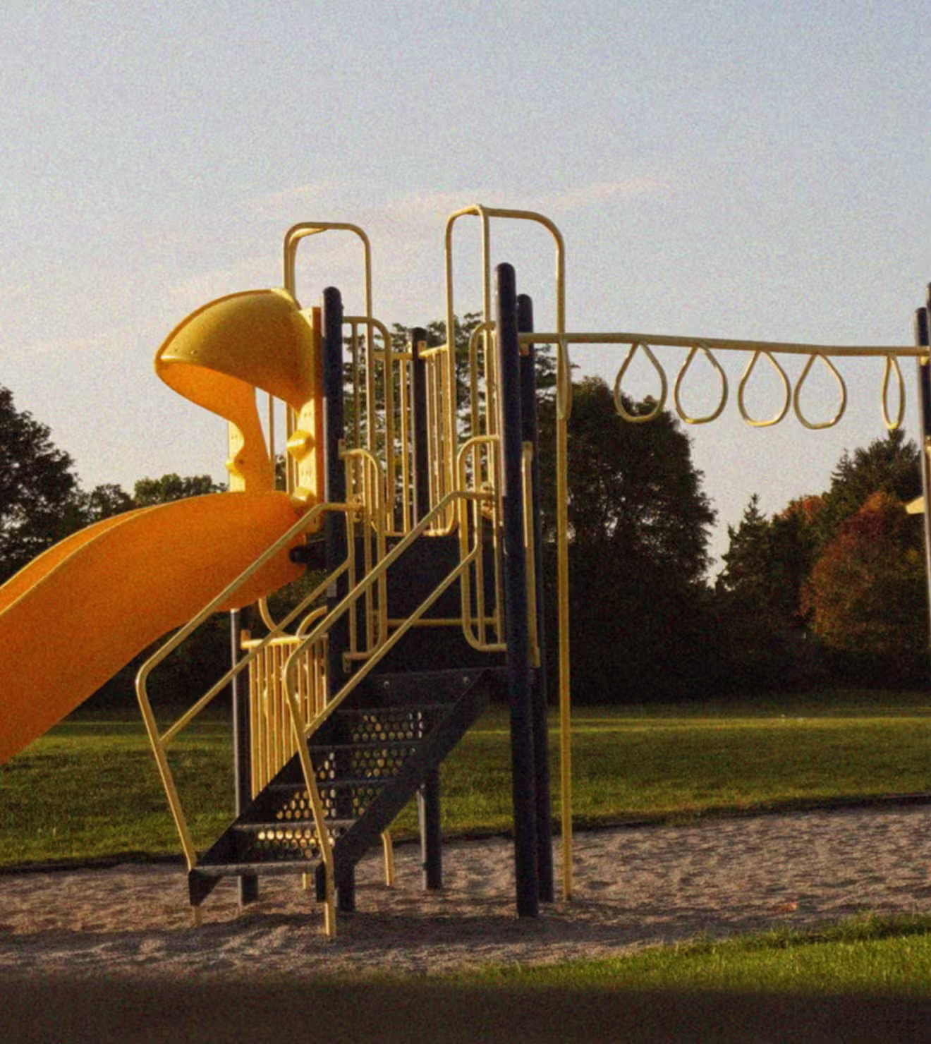 Empty yellow and black playground structure with slide and climbing rings on a grassy field under a clear sky.