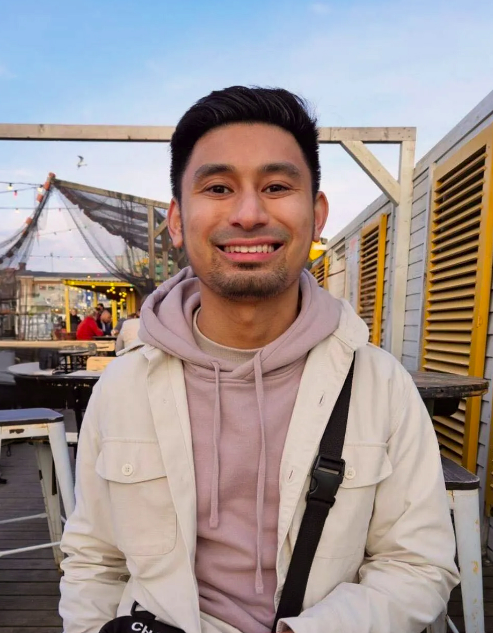 Smiling man wearing a light beige jacket and pink hoodie standing outdoors near tables and yellow shutters.