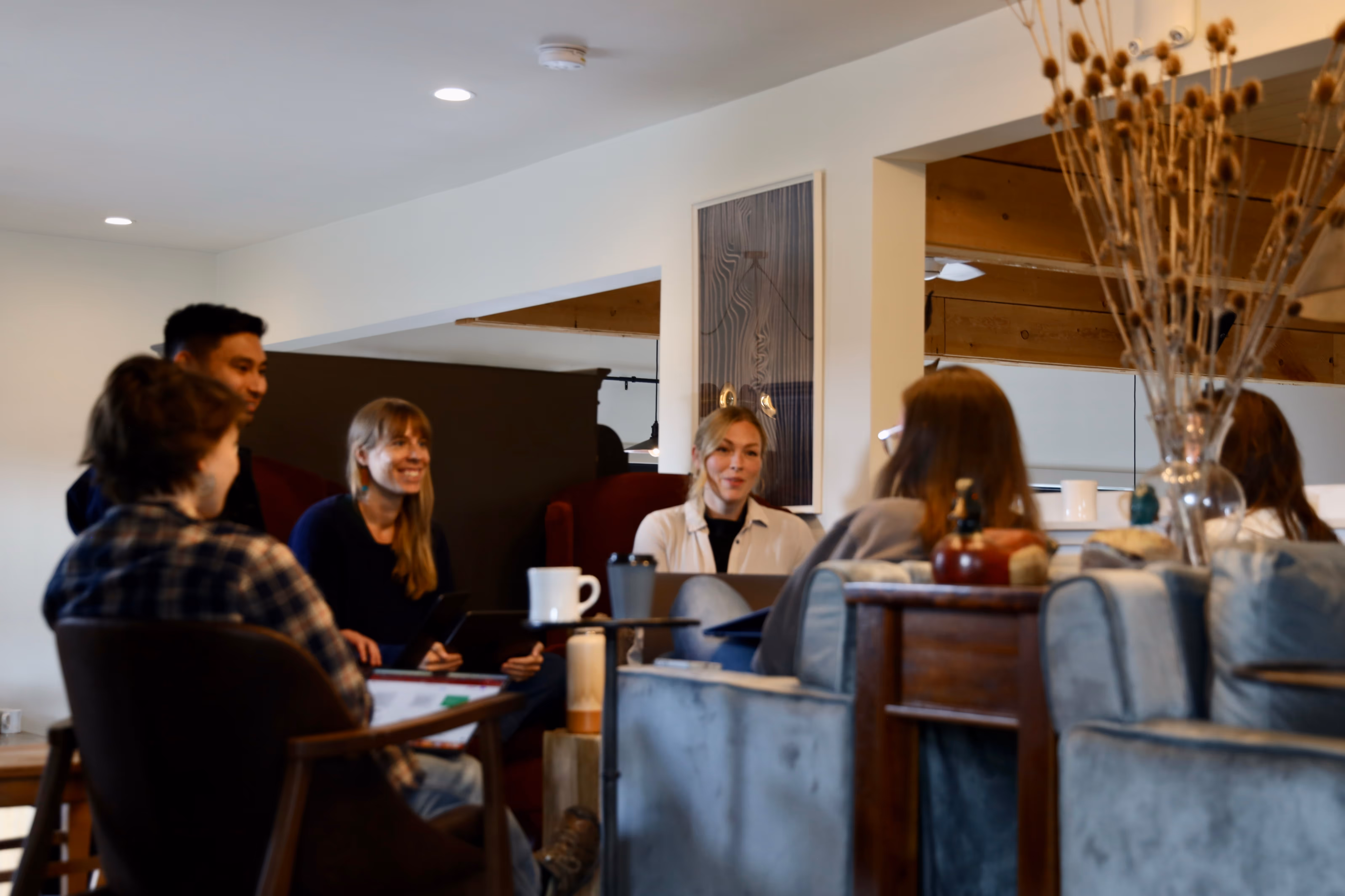 Group of five people sitting in a cozy living room having a discussion, with coffee mugs and laptops nearby.