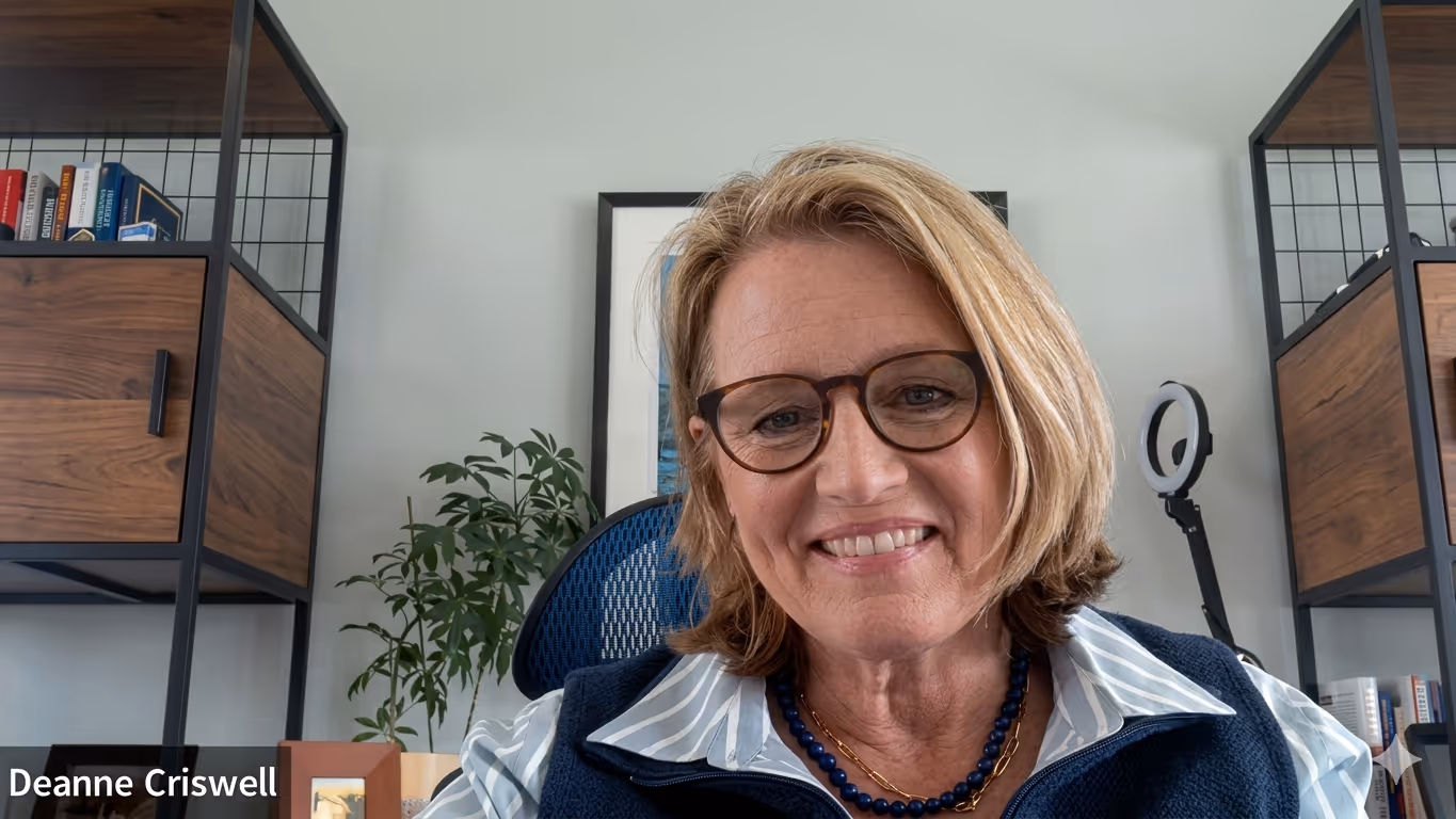 Smiling woman with glasses and layered blonde hair sitting in a modern office with wooden shelves and a ring light in the background.