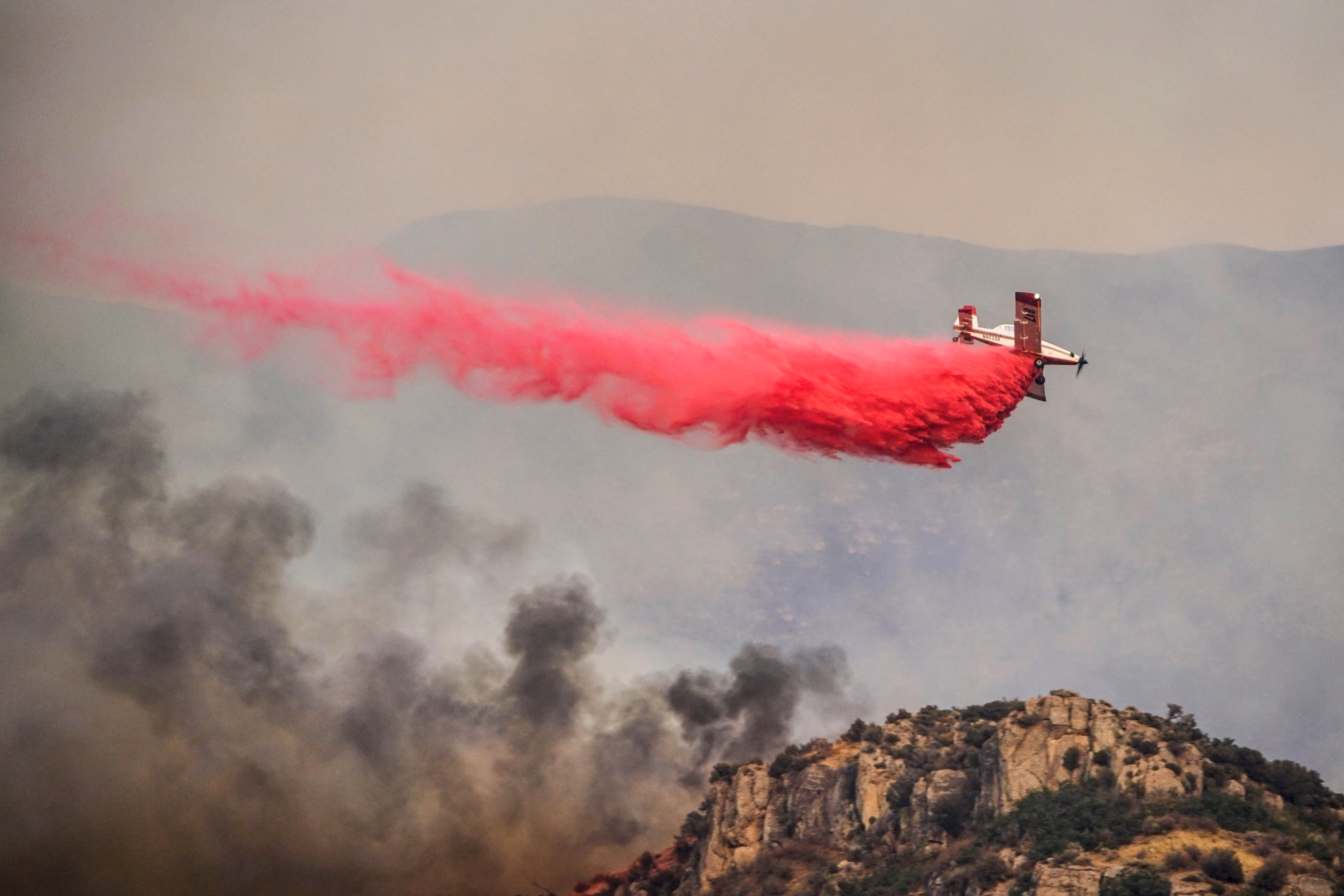 Airplane dropping red fire retardant over smoky wildfire near rocky hill under hazy sky.