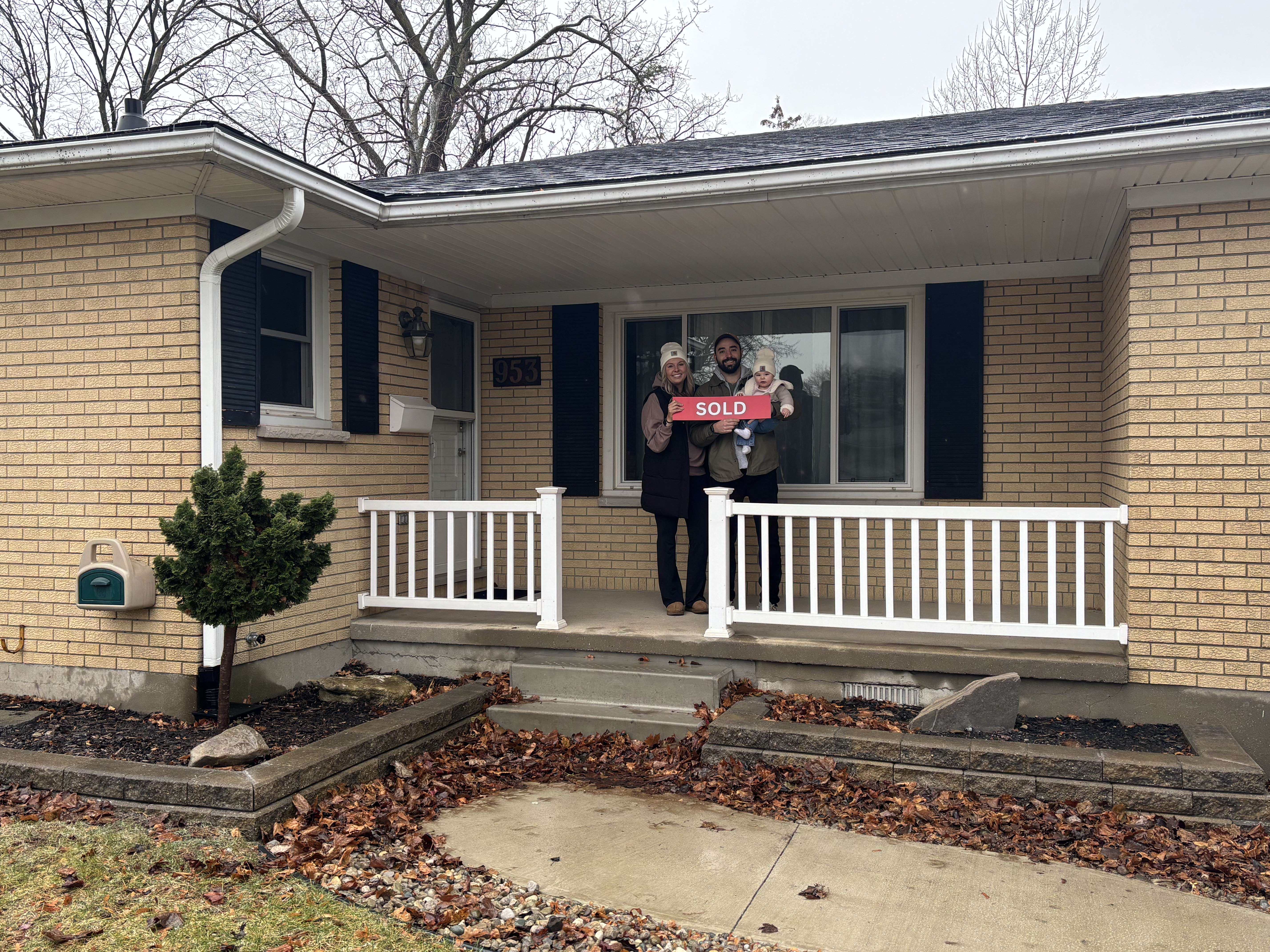 Happy family of three standing on the porch of a beige brick house holding a red SOLD sign.