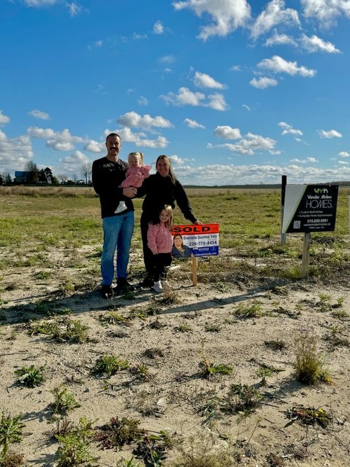 Family of four standing on a vacant lot with a sold real estate sign under a blue sky with scattered clouds.