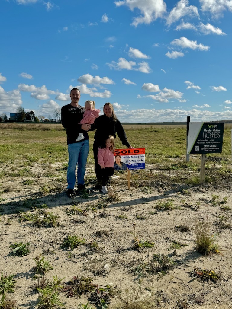 Family of four standing on a vacant lot with a sold real estate sign under a blue sky with scattered clouds.