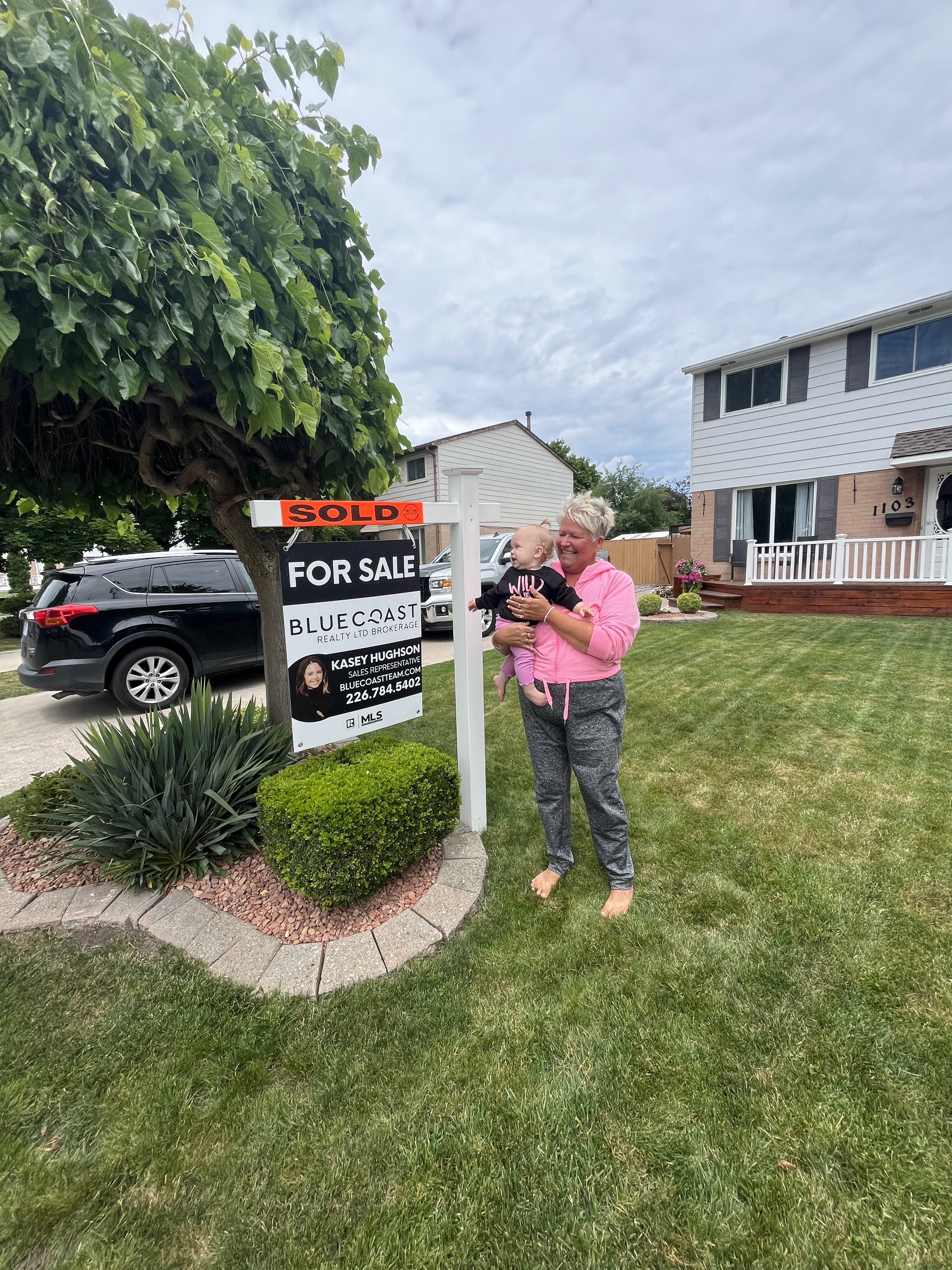 Woman in pink hoodie holding a baby stands on a lawn next to a sold real estate sign in front of a two-story house.