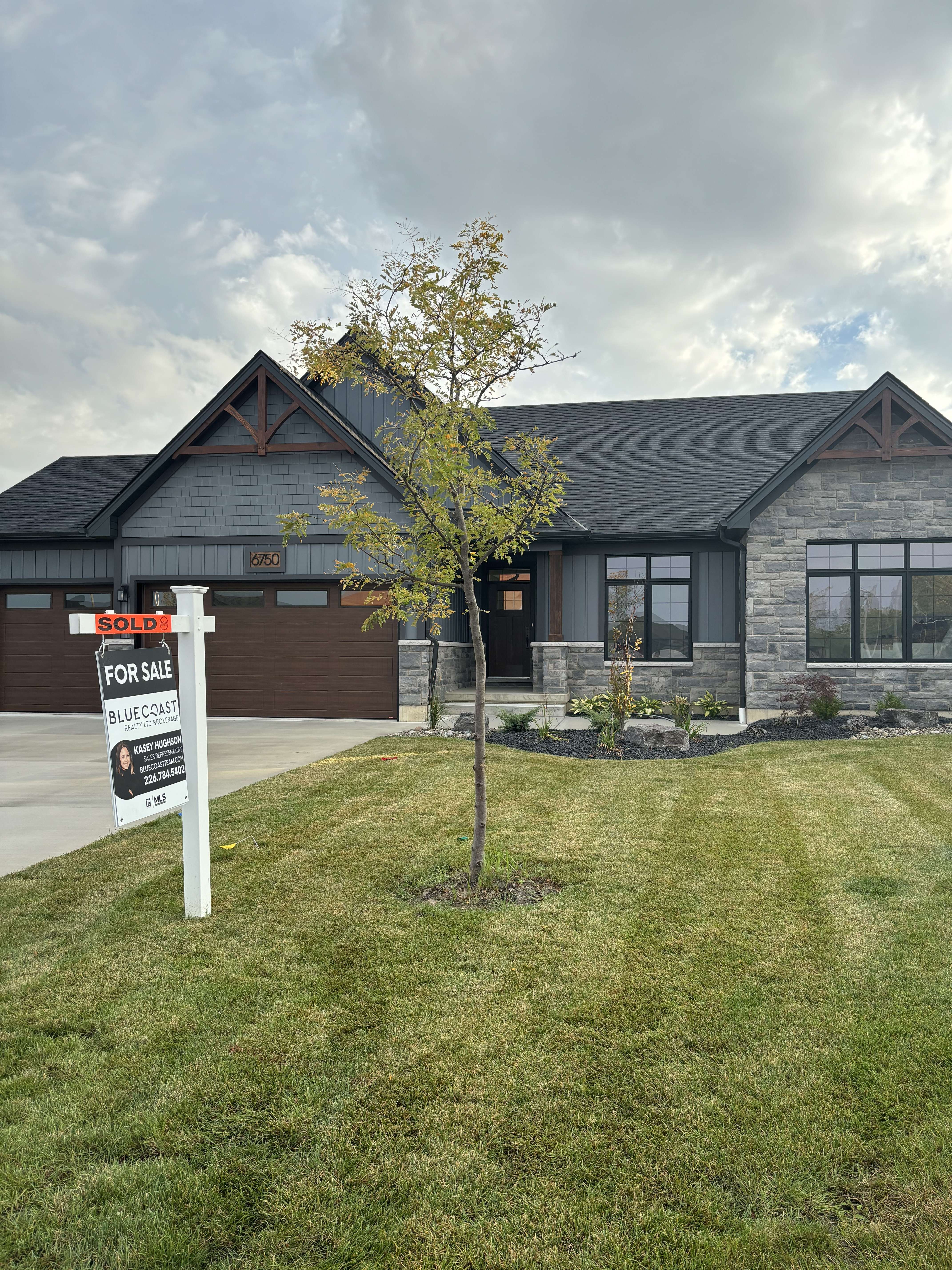 Single-story modern house with a stone and siding exterior, a tree in the front yard, and a sold real estate sign.