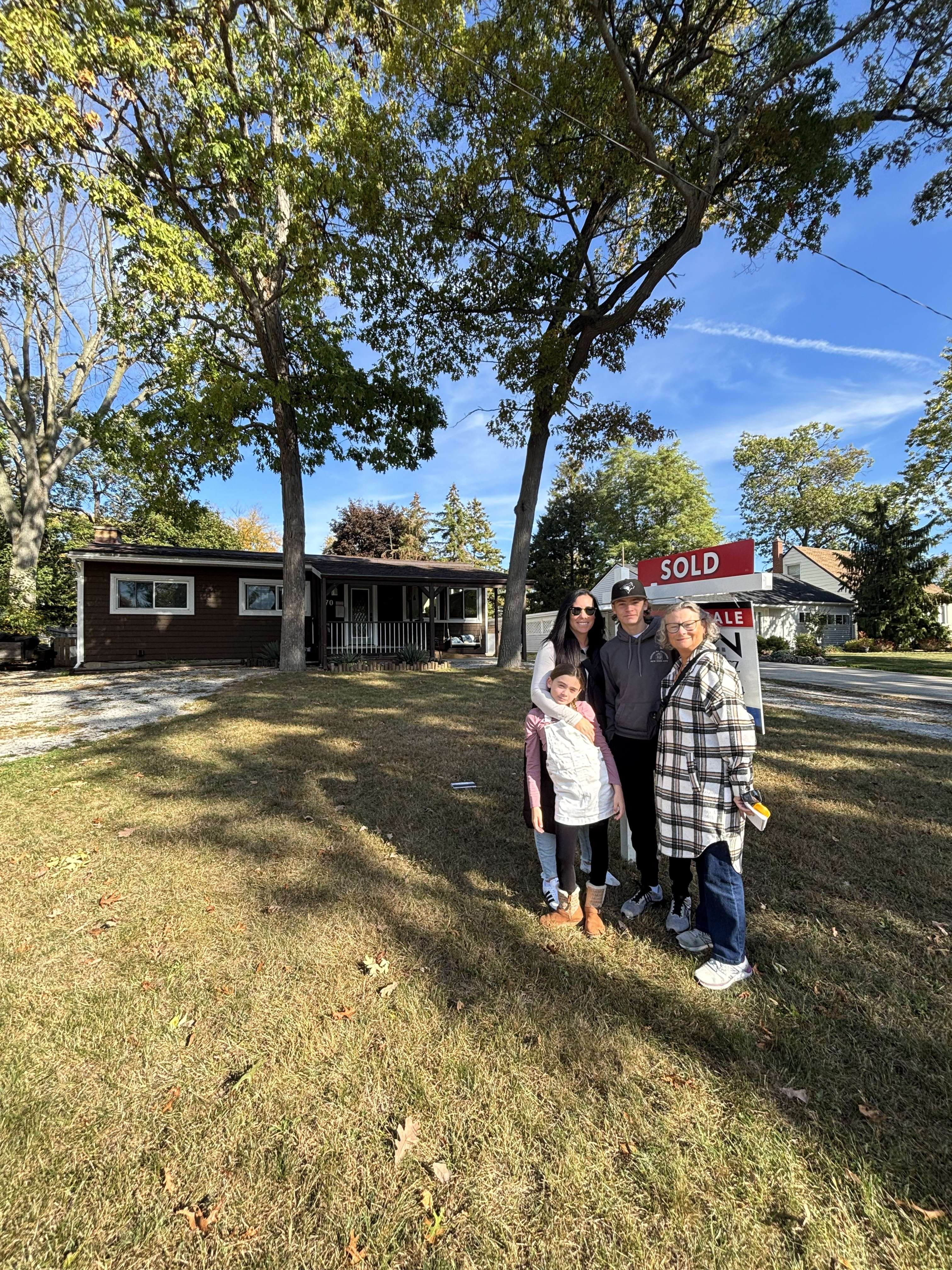 Four people standing on a lawn in front of a house with a large Sold real estate sign.