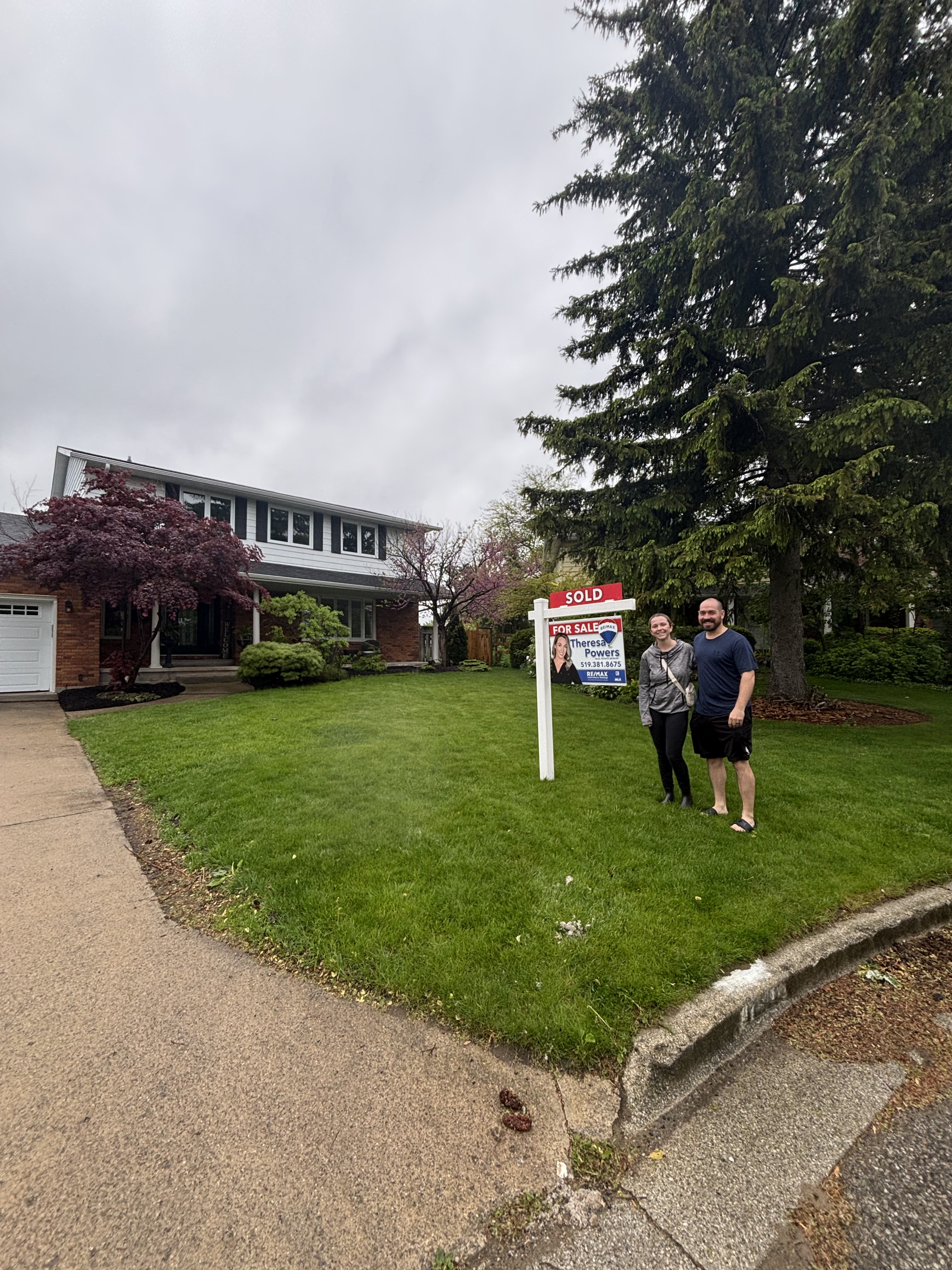 Smiling couple standing beside a sold sign on the lawn of a two-story suburban house.