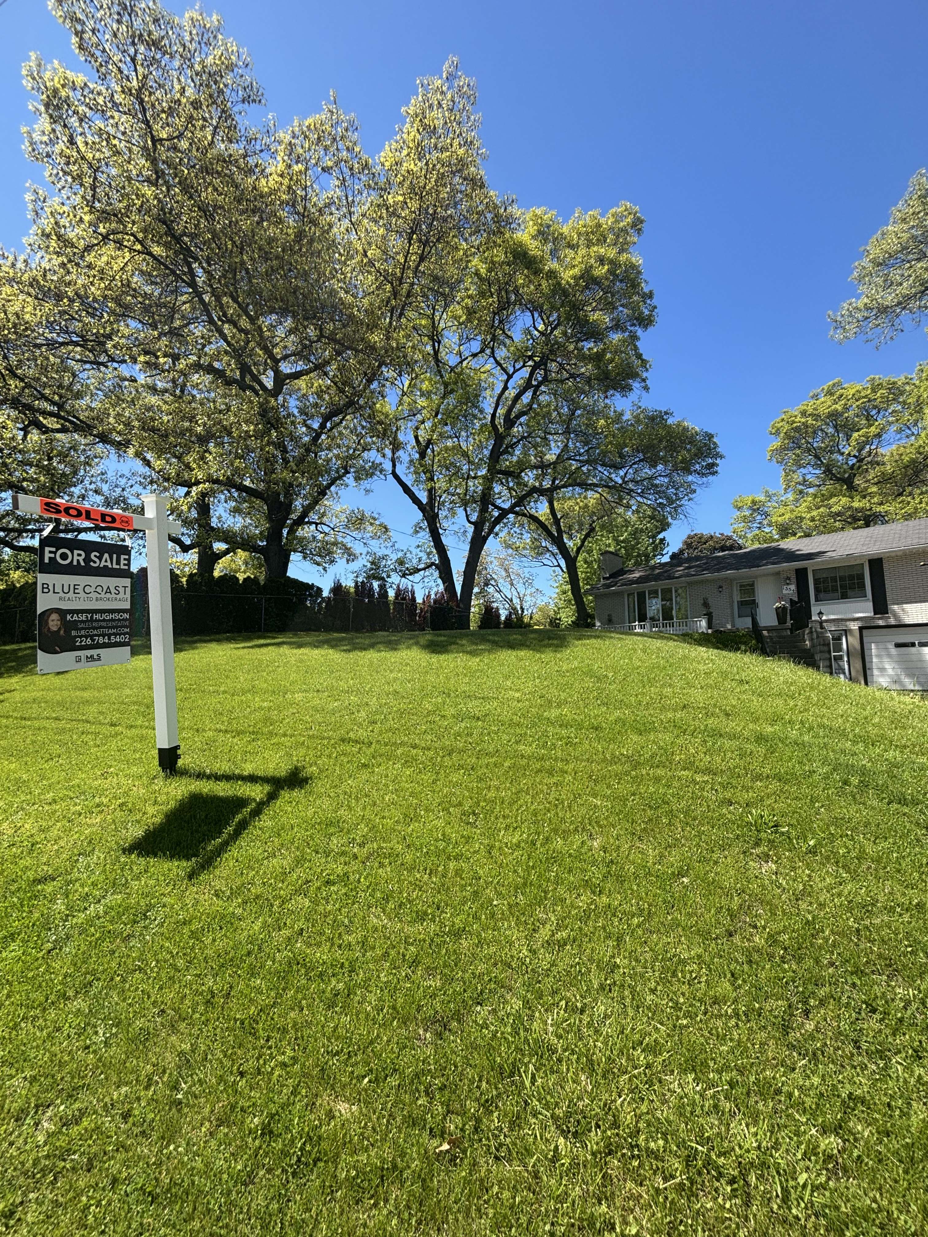 Grassy front yard with a 'Sold' and 'For Sale' real estate sign, large trees, and a white house on a sunny day.