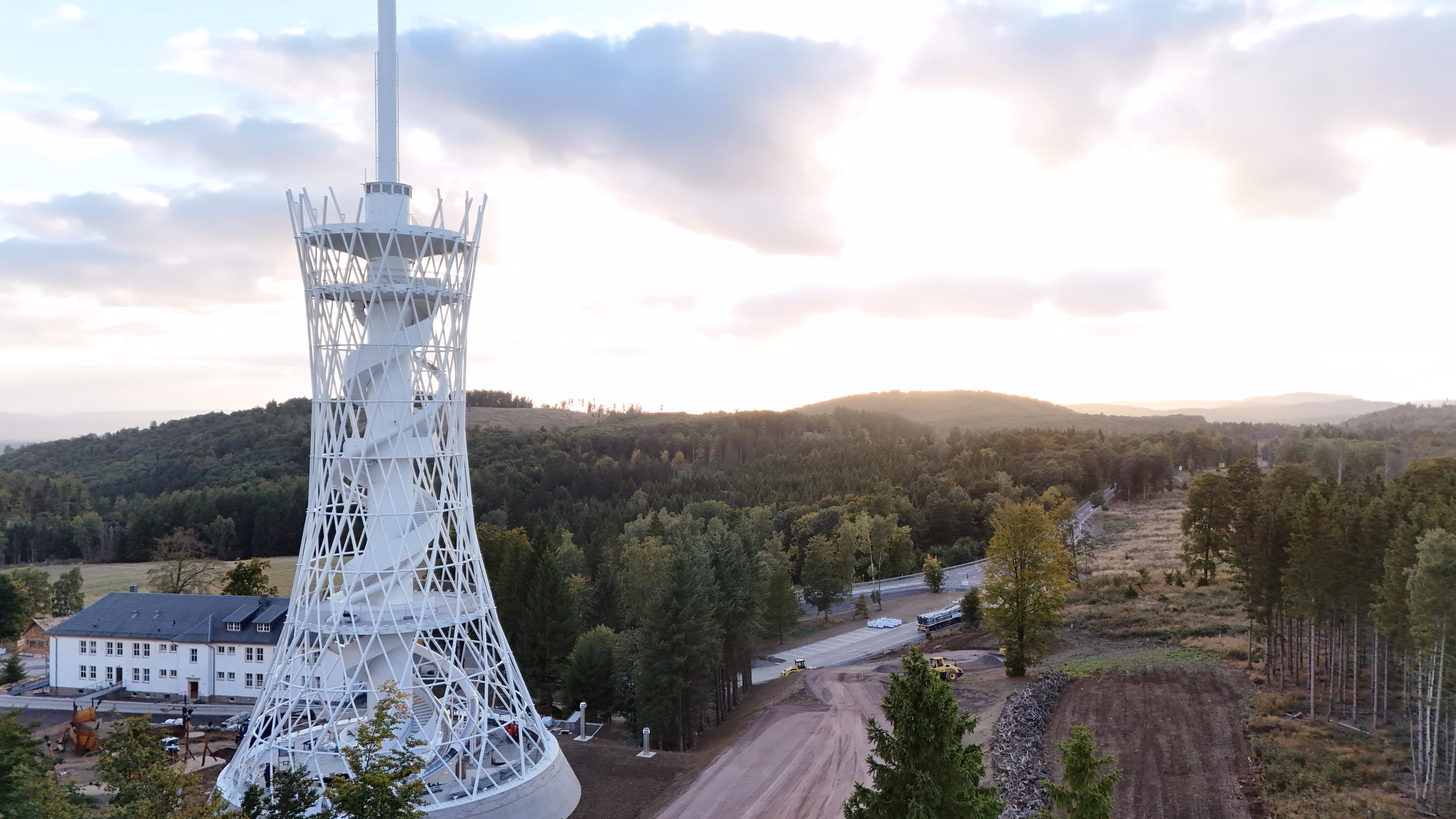 Witte uitkijktoren met spiraaltrap omgeven door bos en een landweg bij zonsondergang.