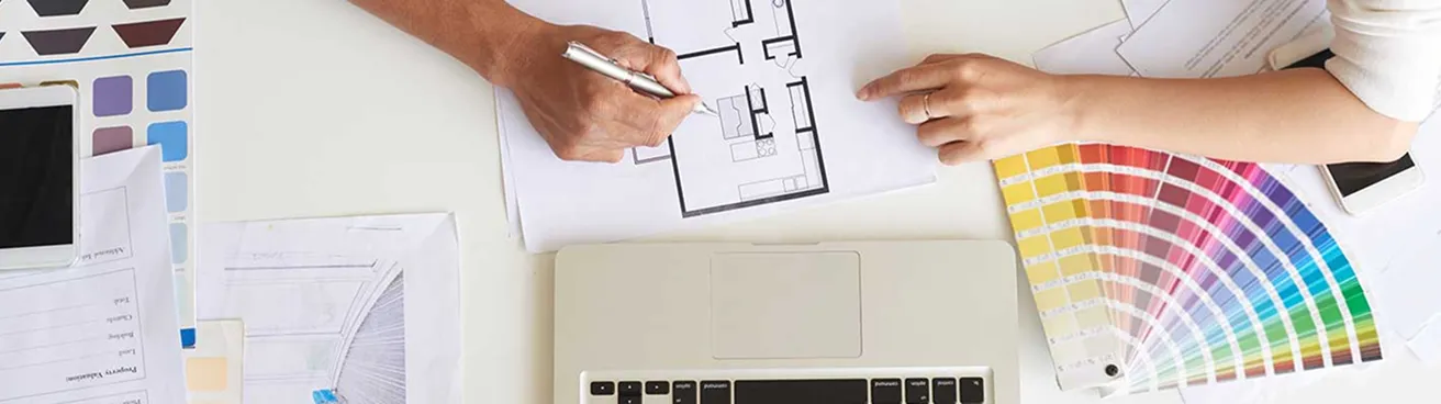 Two people reviewing an architectural floor plan with a laptop, color swatches, and paperwork on a desk.