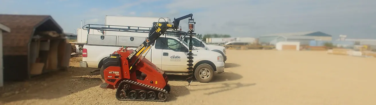 Red Ditch Witch SK755 mini skid steer with auger attachment next to white utility trucks on a gravel lot.