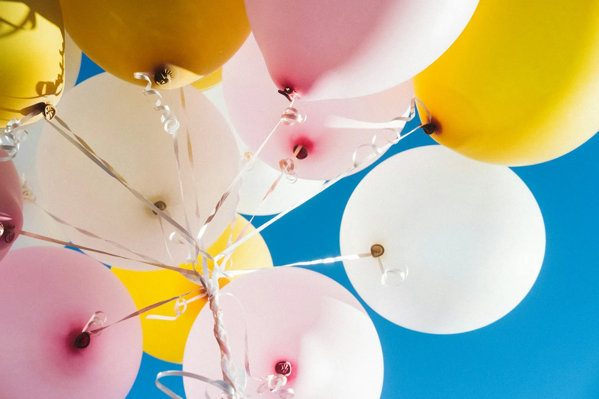 Cluster of yellow, pink, and white balloons with curling ribbons against a clear blue sky.
