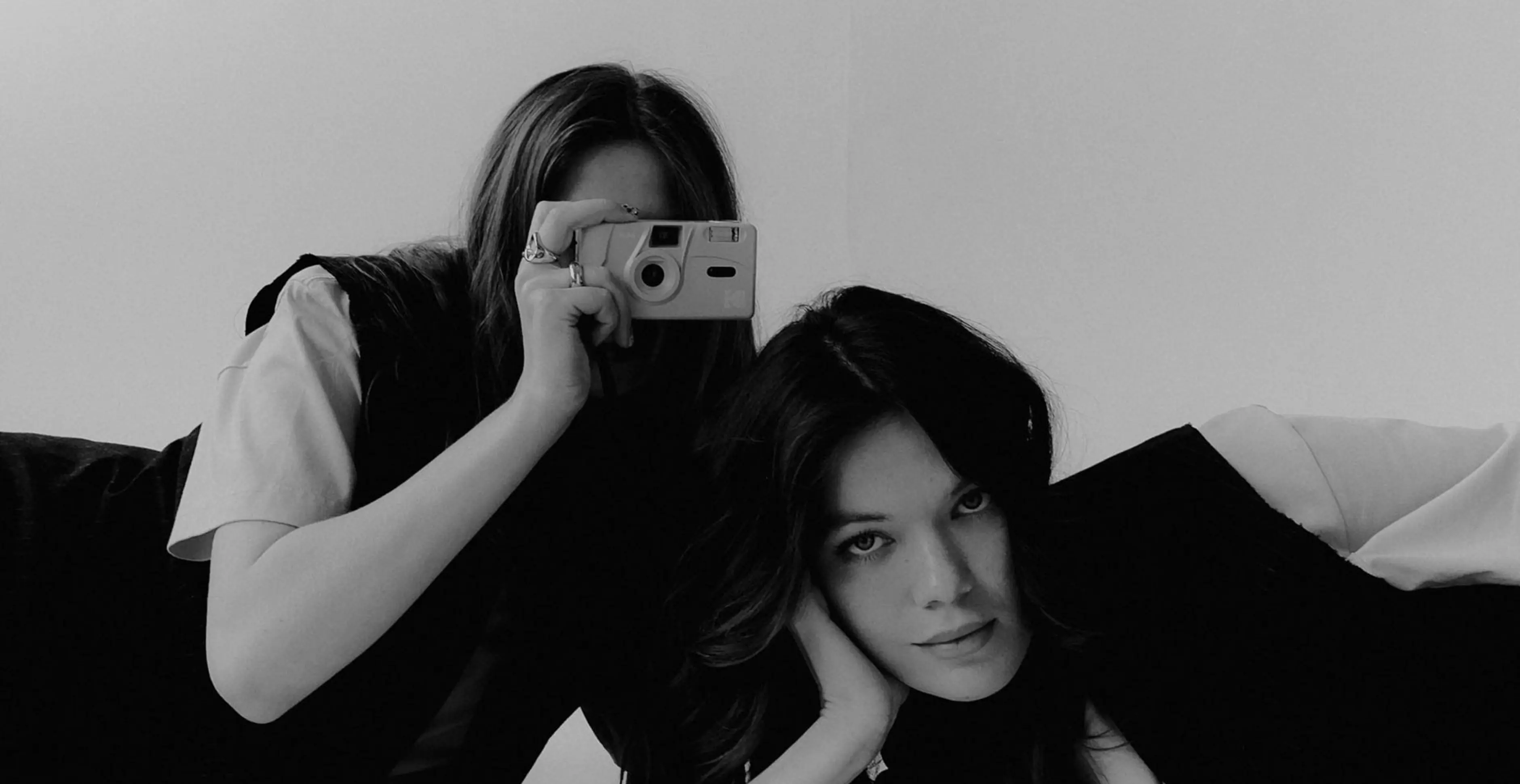 FANDS MARKETING AGENCY —  Black and white photo of two women, one holding a camera up to her face while the other rests her head on her hand looking at the camera.