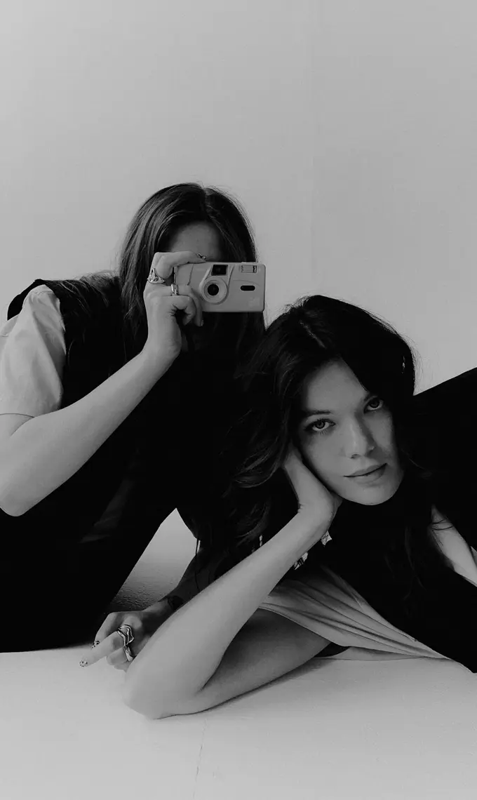 FANDS MARKETING AGENCY —  Black and white photo of two women; one is lying on her side looking at the camera, the other is holding a camera up to her face to take a picture.