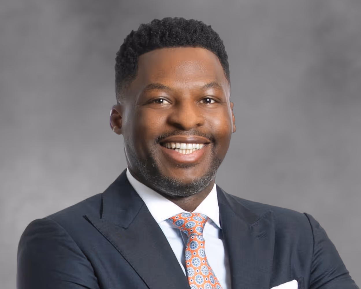 J smiles while wearing a a dark suit with a patterned orange tie and white pocket square, arms crossed against a gray background.