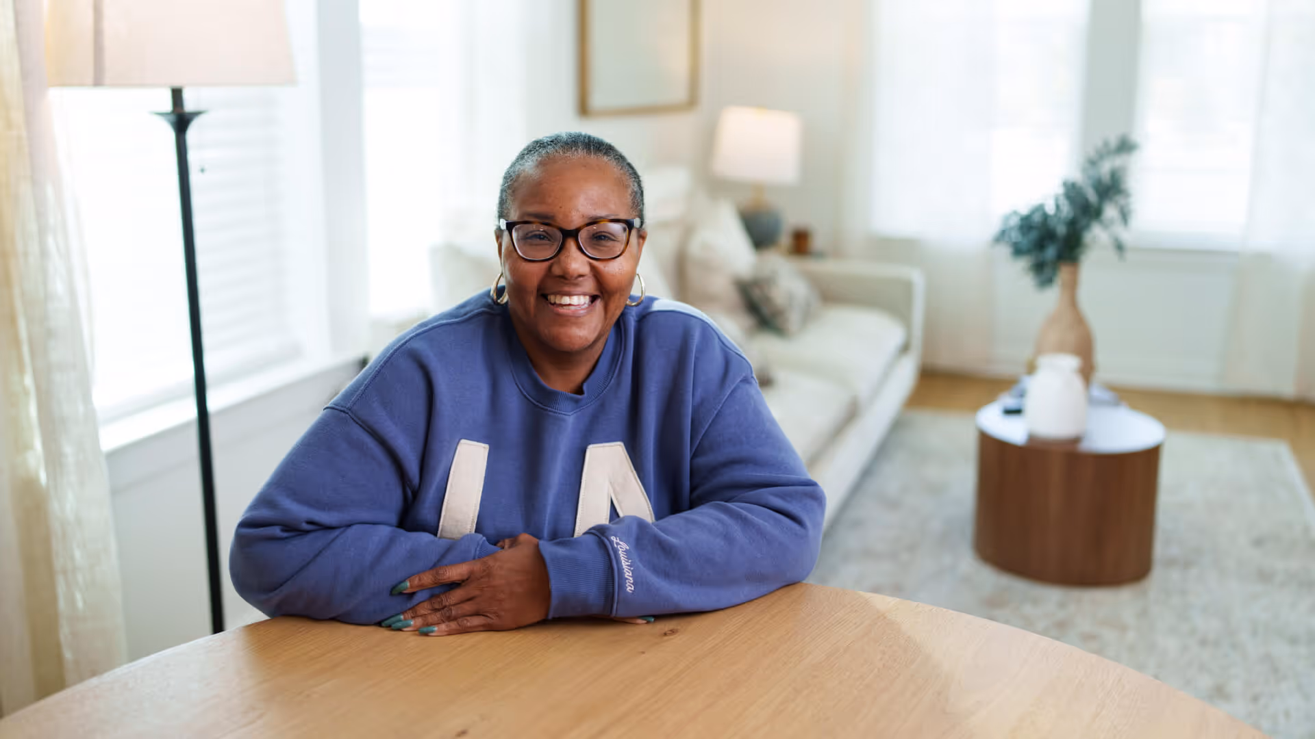 A smiling woman in a blue "LA" sweatshirt sits at a wooden table in a bright, modern living room.