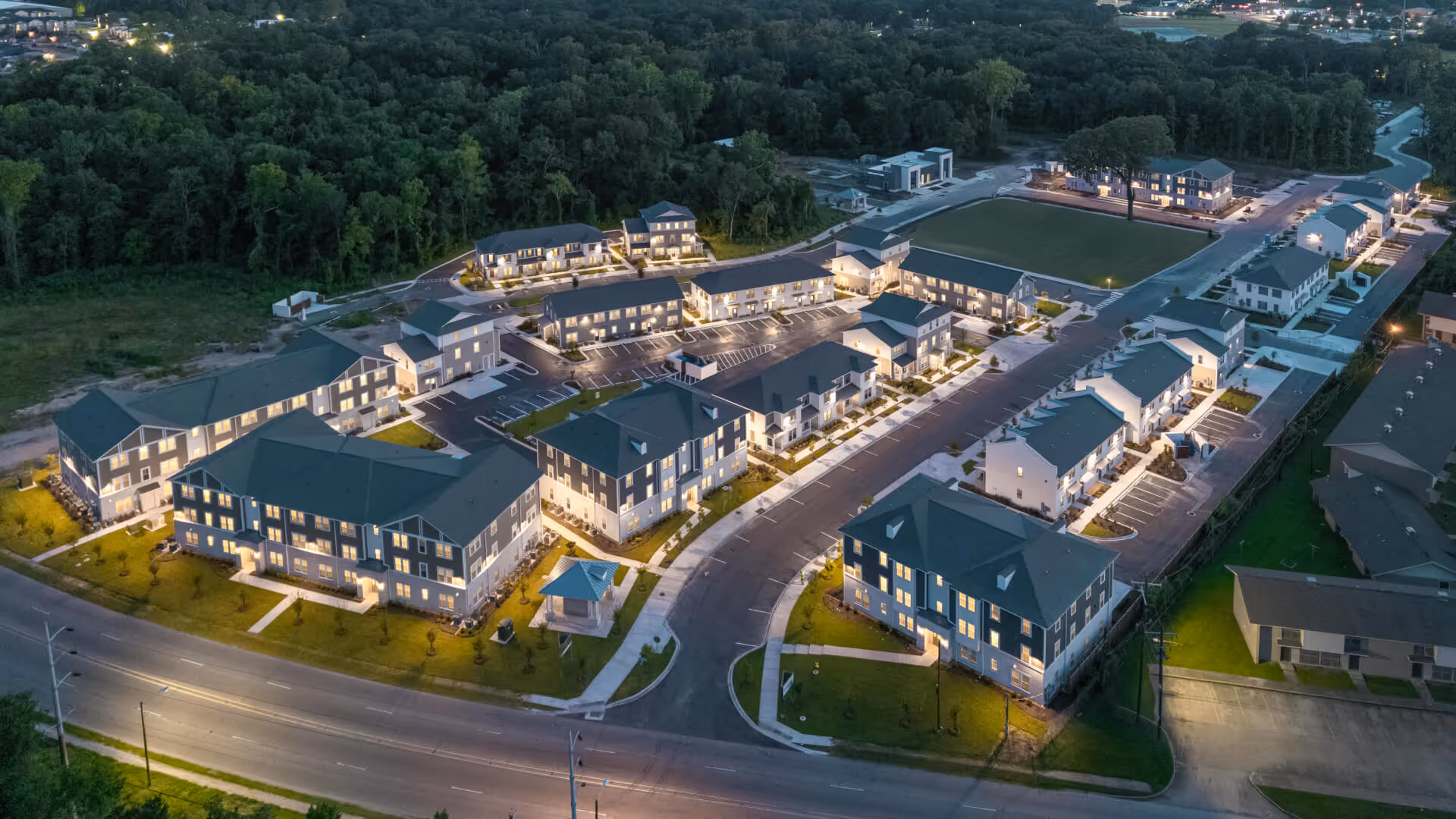 Aerial view of a modern apartment complex at sunset with glowing lights and a golden horizon.