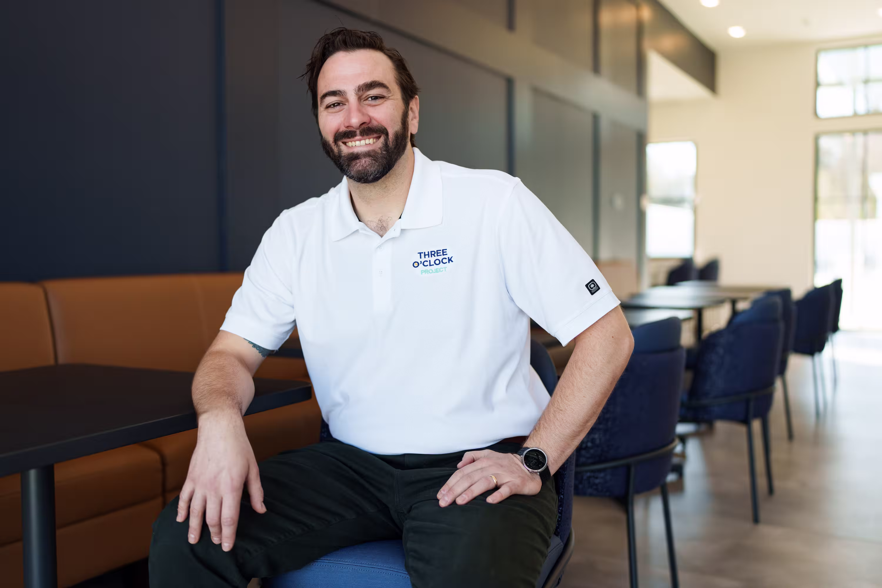A man in a white "Three O'Clock Project" polo shirt sits and smiles in a modern cafeteria or lounge.