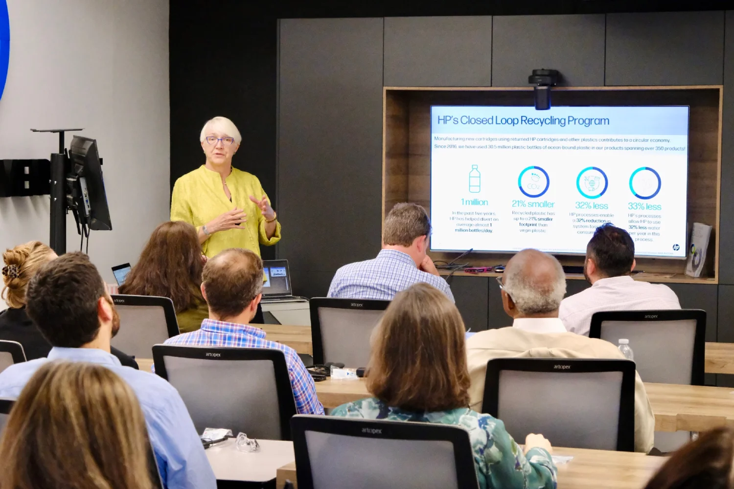 Woman in yellow blouse giving a presentation about HP's Closed Loop Recycling Program to an audience in a conference room.