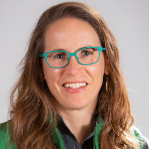 Smiling woman with long light brown hair wearing green glasses and a green jacket against a light background.