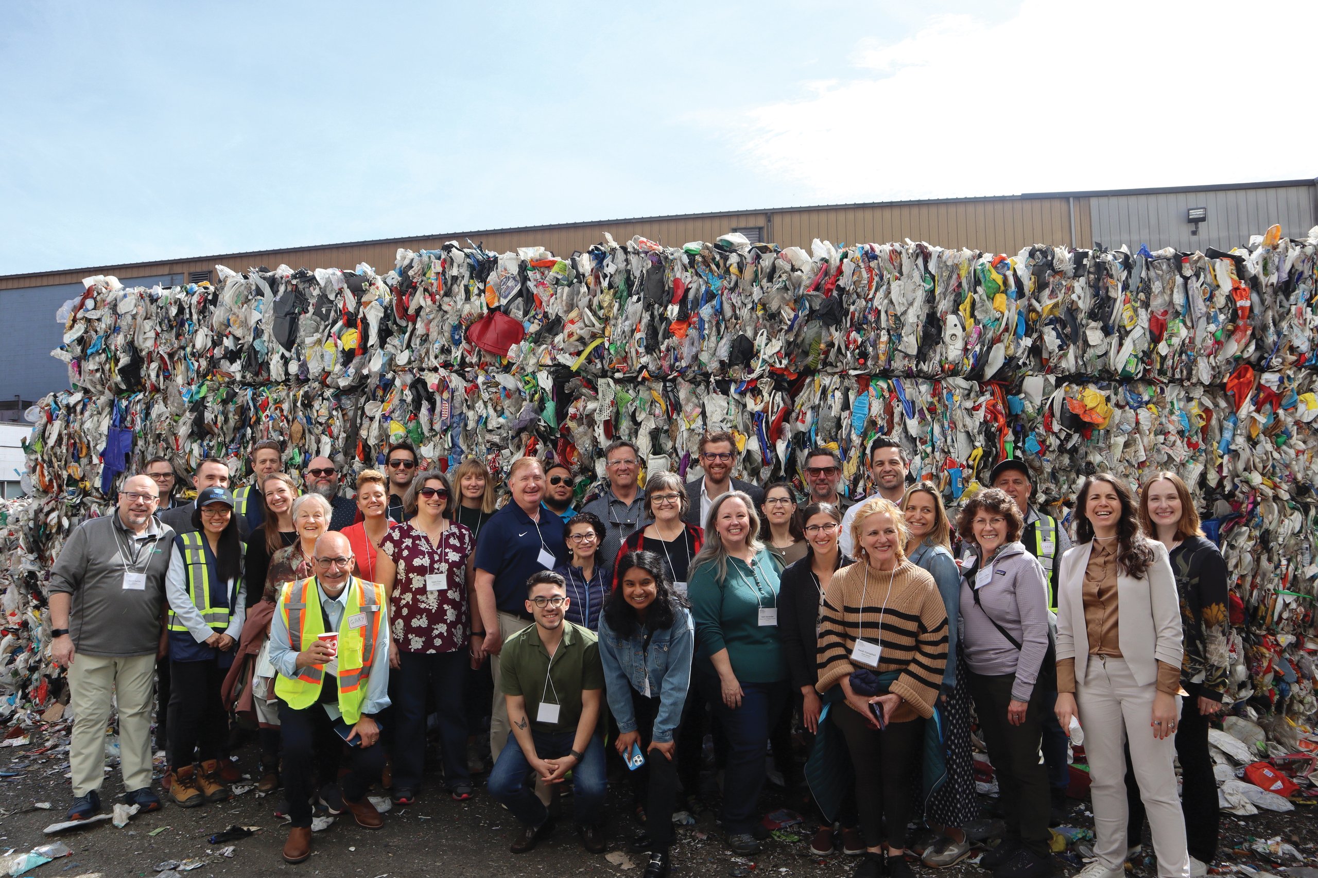 Group of diverse adults posing in front of large bales of compacted recycled plastic waste outdoors.