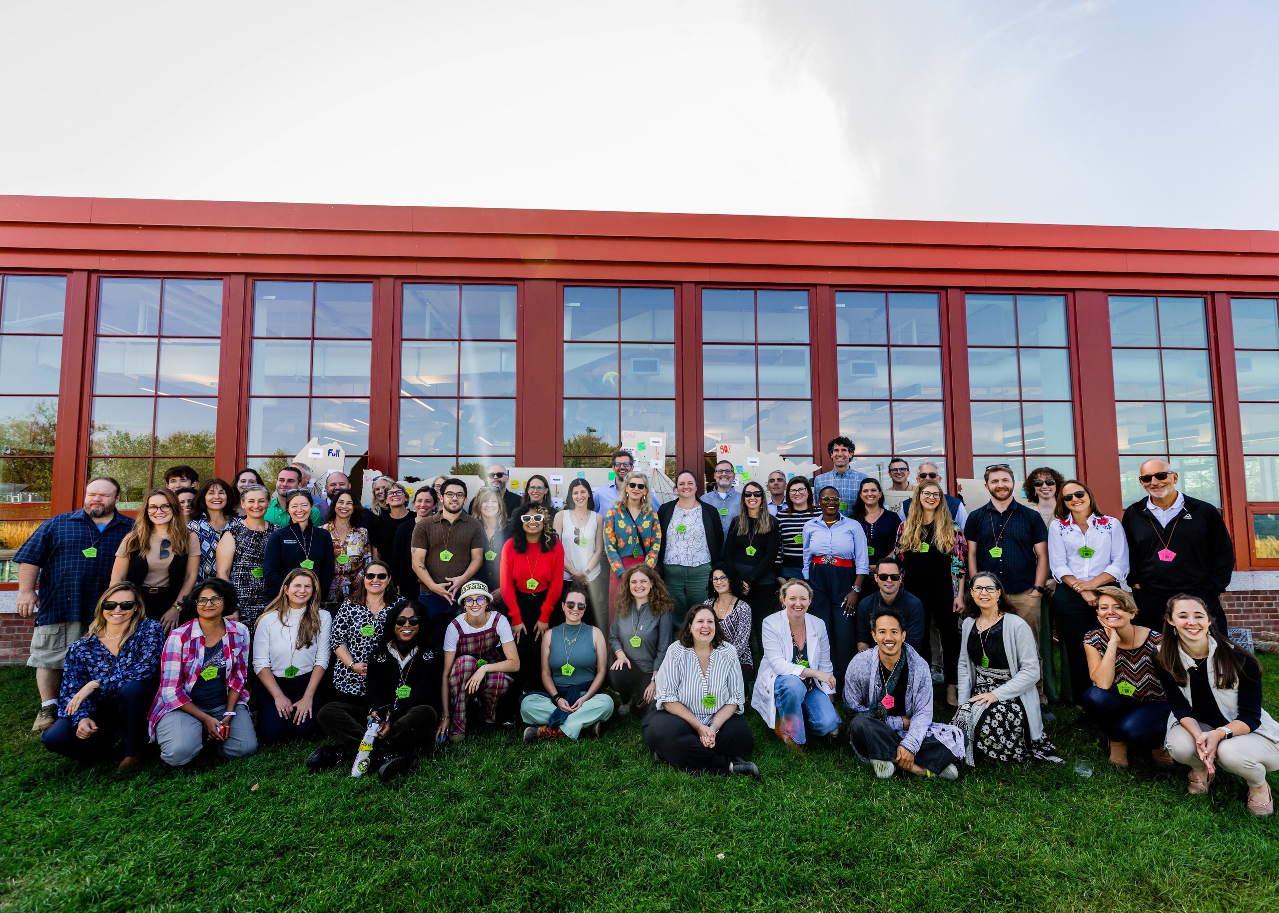 Large diverse group of people posing outside on green grass in front of a building with large red-framed windows under a blue sky.