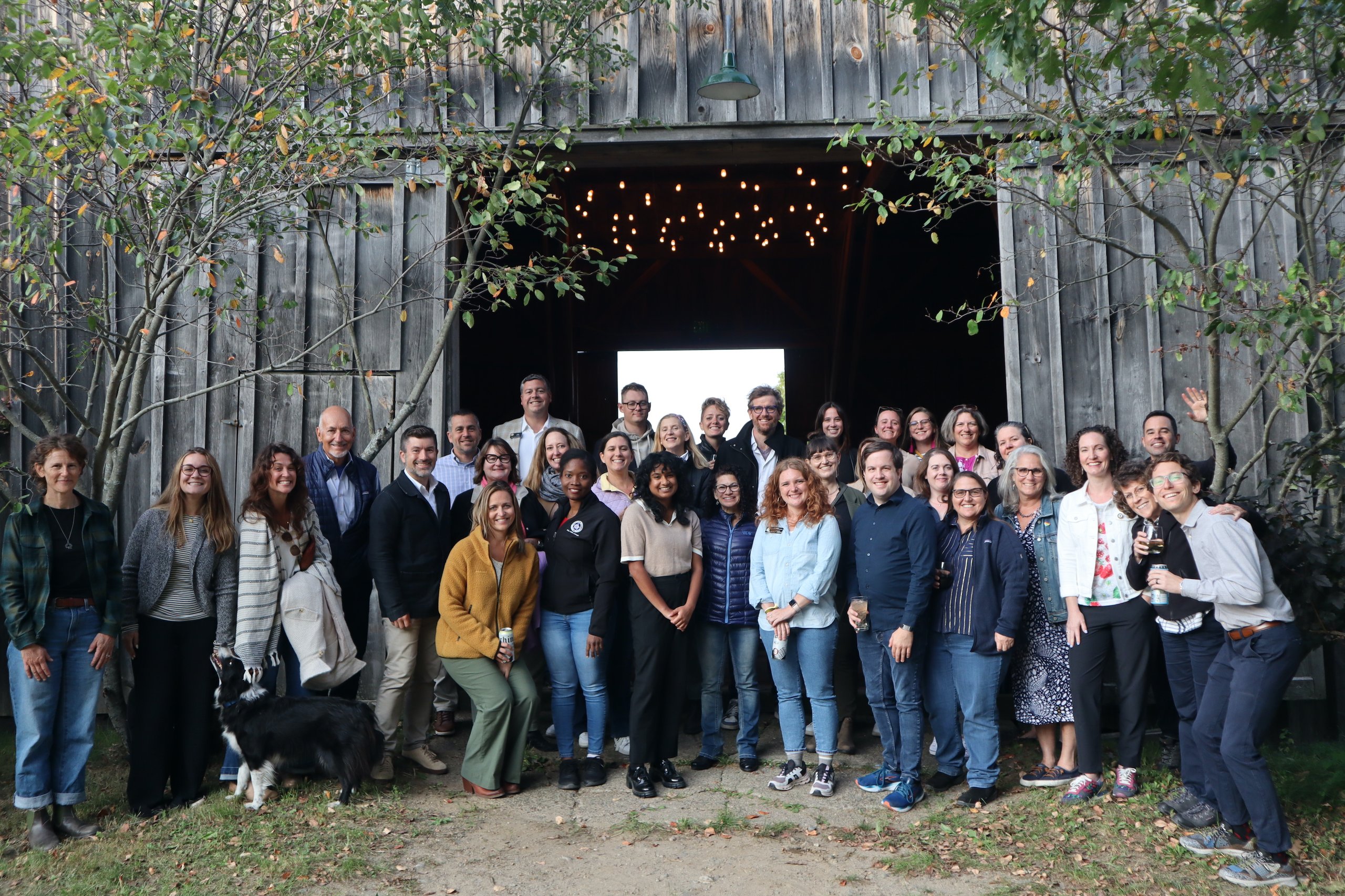 Group of 27 diverse adults standing and smiling in front of a large rustic wooden barn entrance with string lights inside, with a black and white dog on the left.