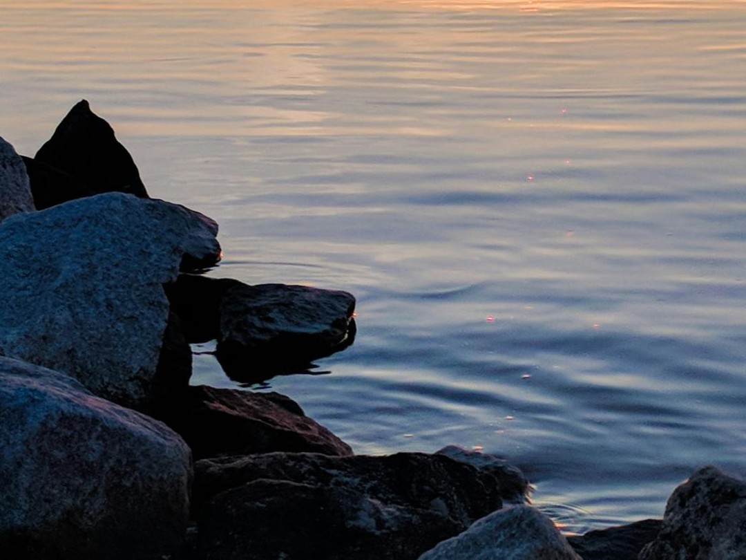 Rocks along the edge of a calm body of water during sunset with soft orange and blue reflections.