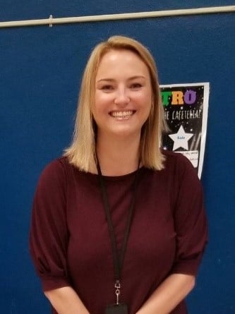 Smiling woman with shoulder-length blonde hair wearing a maroon top and black lanyard standing in front of a blue wall with a colorful poster.