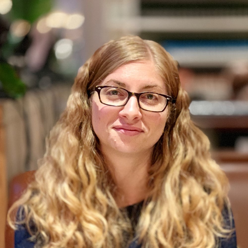 Woman with long wavy blonde hair and glasses smiling softly indoors.