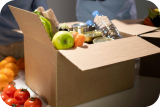 Open cardboard box filled with assorted groceries including fresh produce and canned goods on a table.