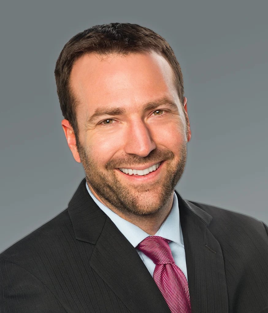 Smiling man with short brown hair and beard wearing a dark suit, light blue shirt, and pink striped tie against a gray background.