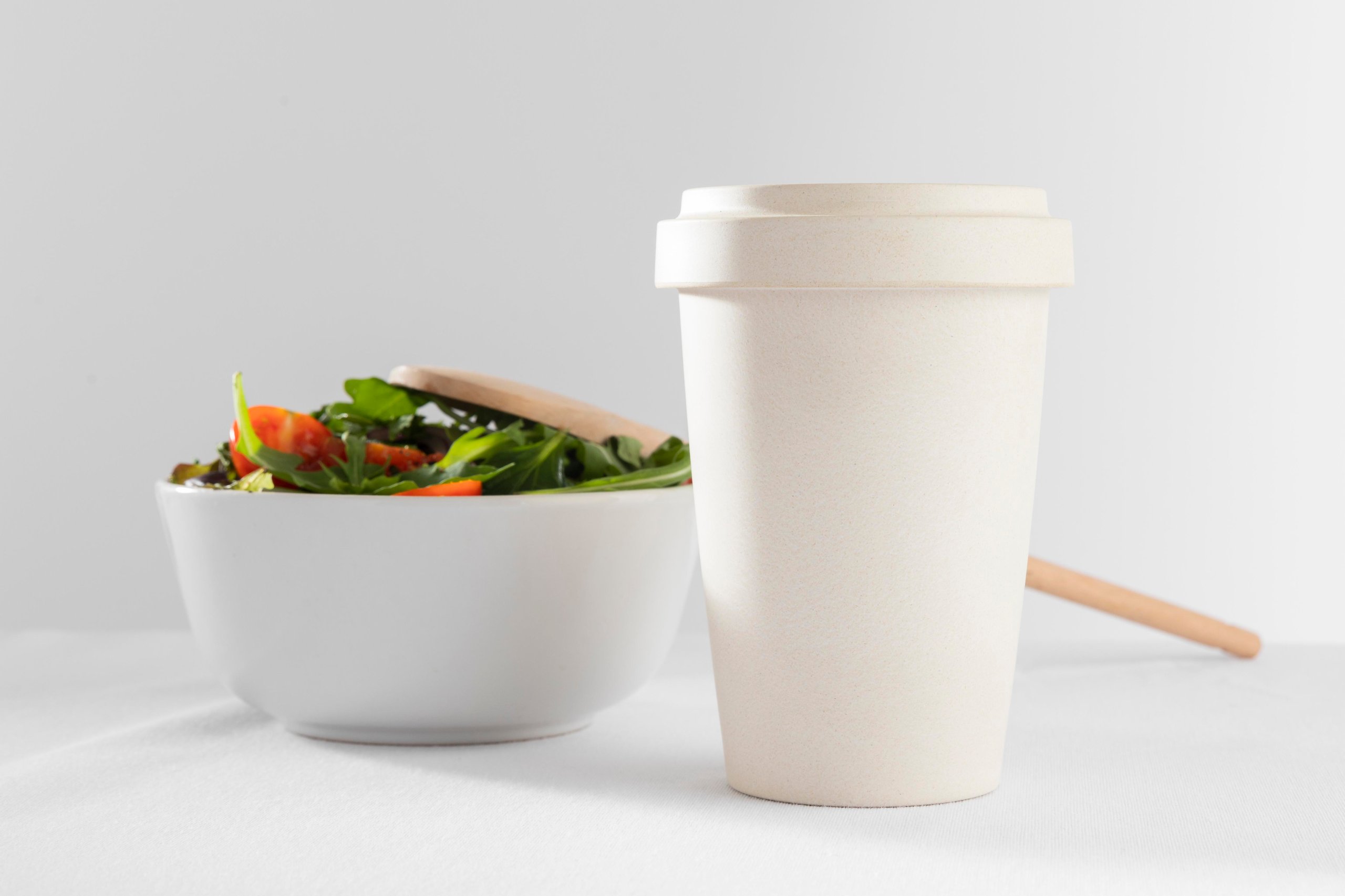 White disposable coffee cup with lid next to a white bowl of fresh salad with leafy greens and tomato slices.