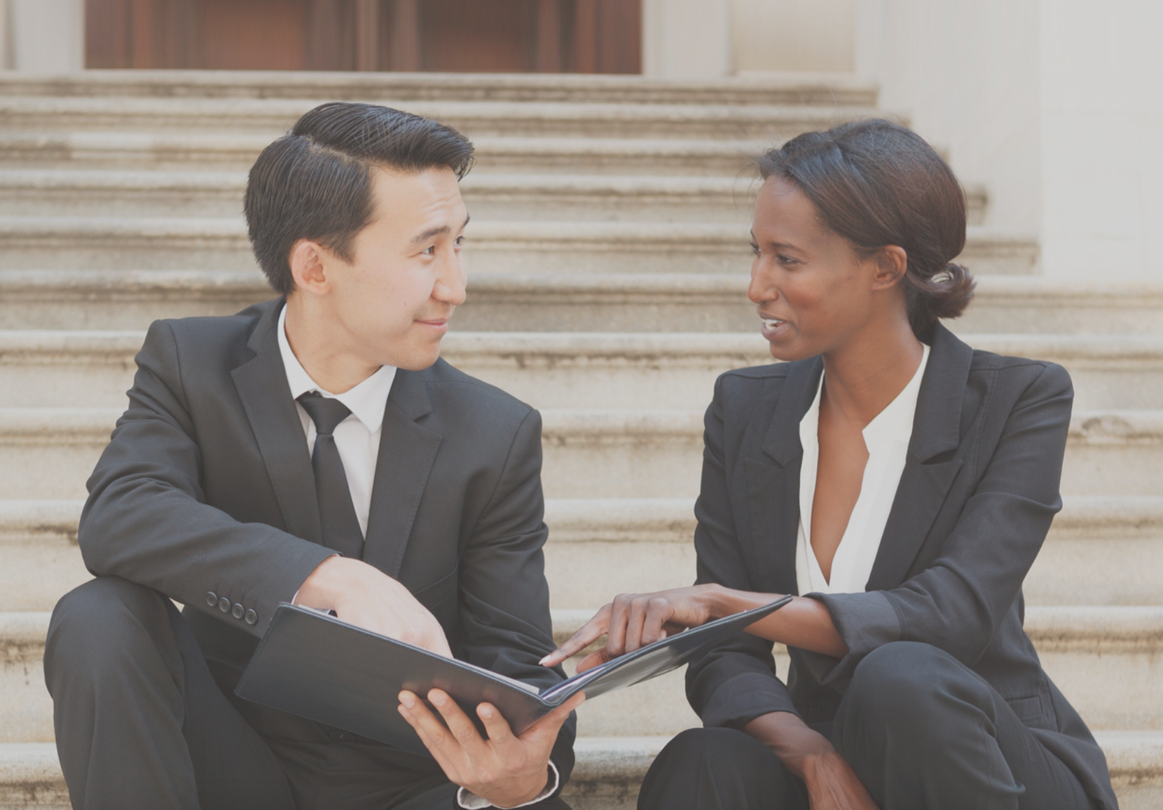 Two business professionals sitting on outdoor steps, discussing content in a folder.