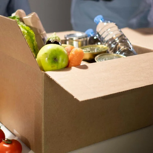 Cardboard box filled with groceries including a green apple, carrots, canned goods, a water bottle, and lettuce.
