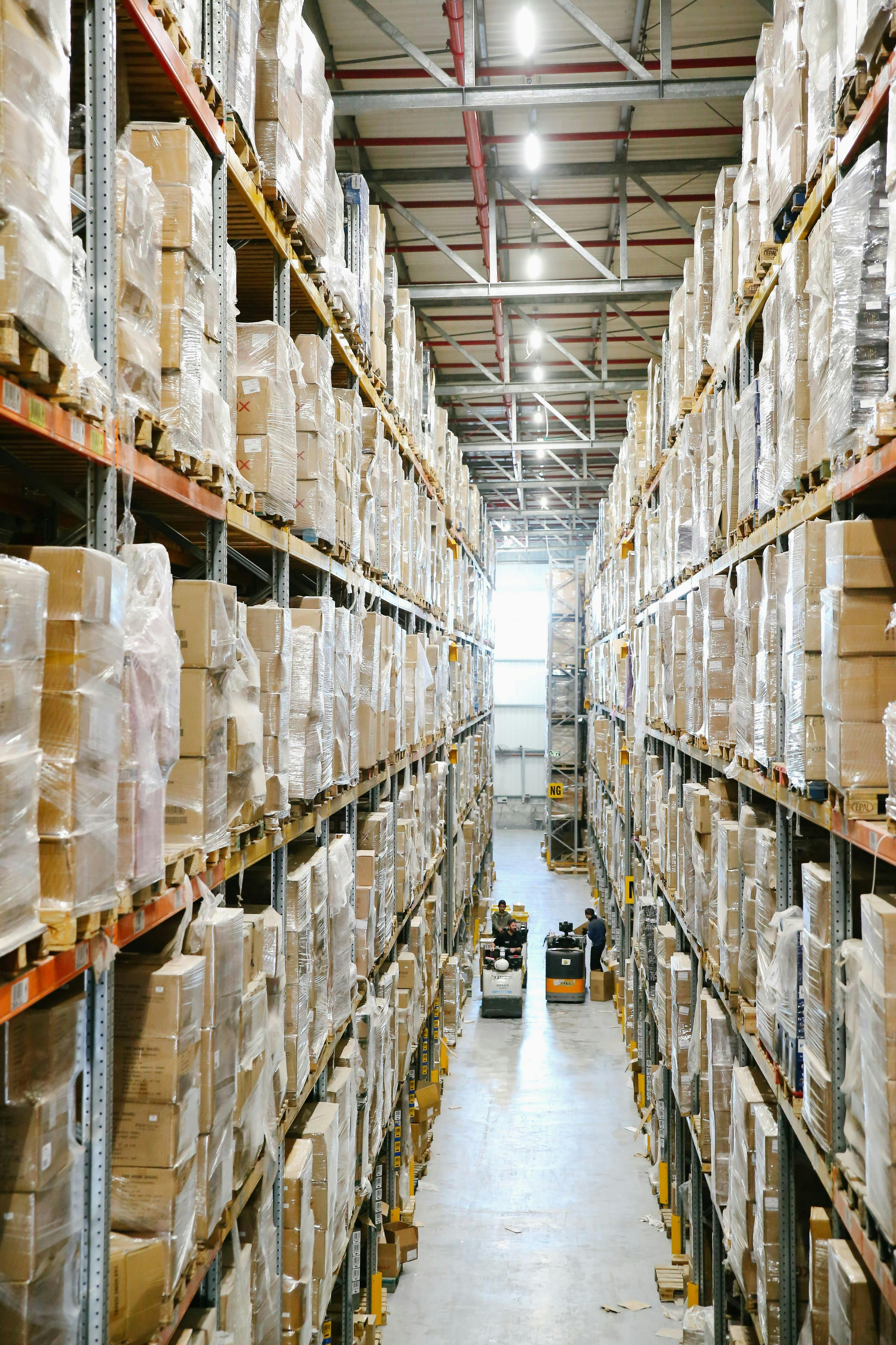 High-ceiling warehouse aisle with tall shelves stacked with plastic-wrapped cardboard boxes and two workers operating forklifts.