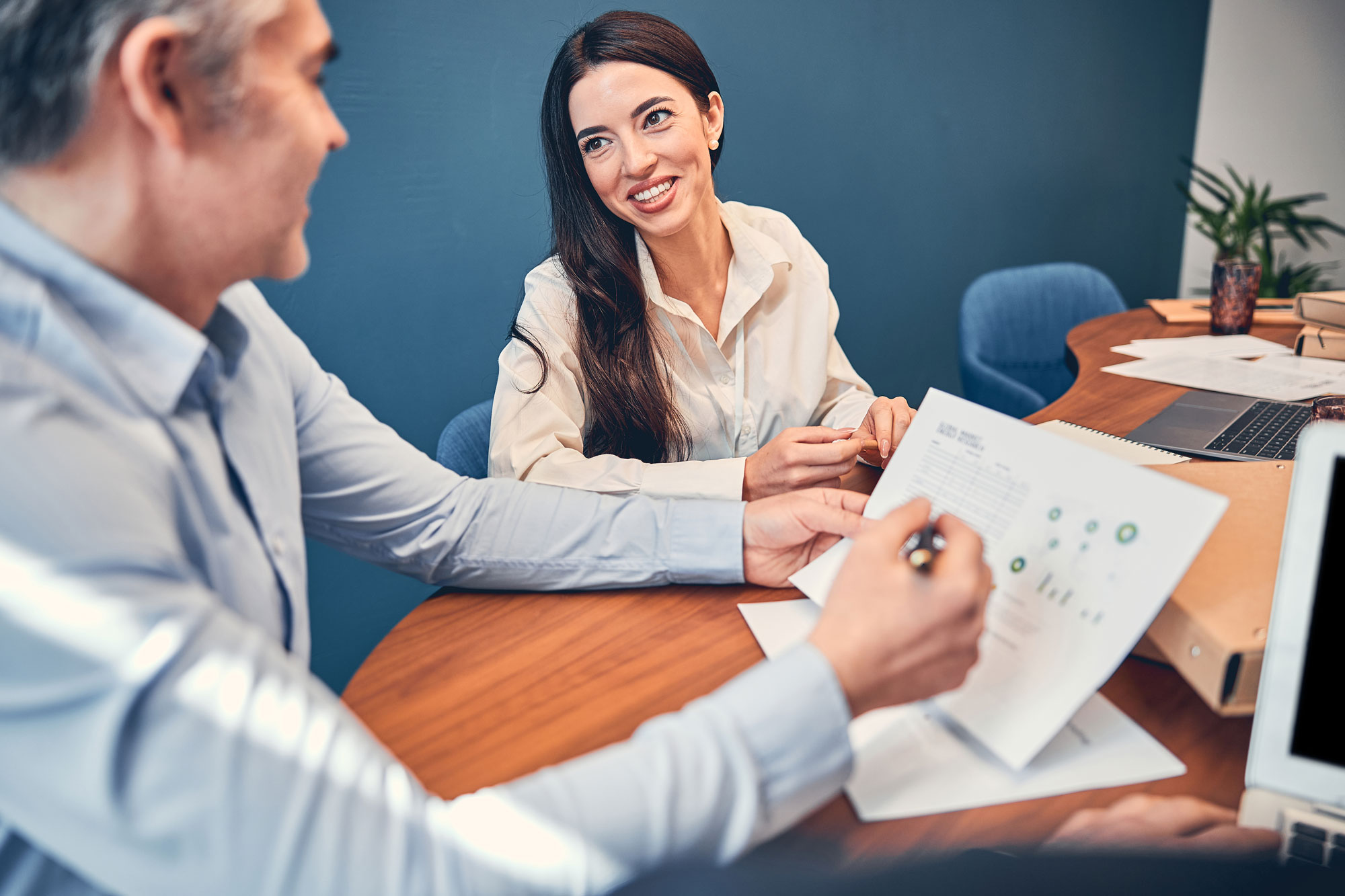 Une femme souriante en chemise blanche discute avec un homme qui lui montre des documents dans un bureau moderne.
