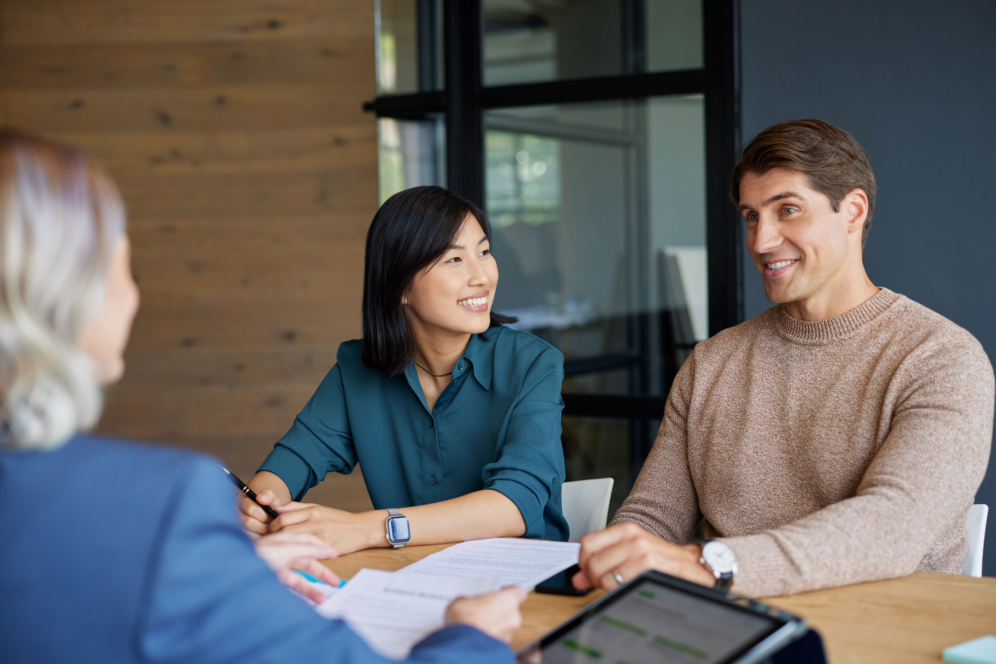 Deux clients souriants en discussion avec une conseillère financière autour d'une table avec des documents et une tablette.