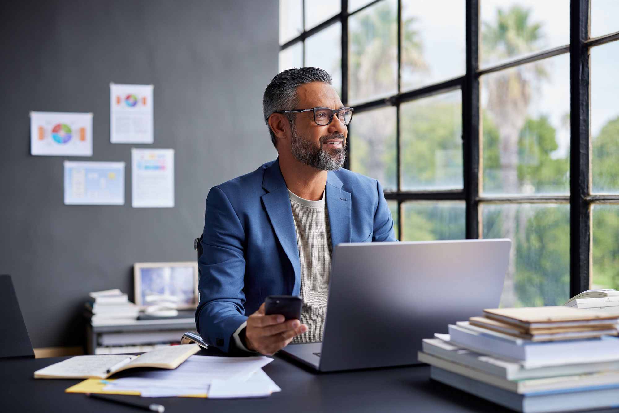Homme d'affaires portant des lunettes et un blazer bleu, assis à un bureau avec un ordinateur portable et un téléphone, regardant par une grande fenêtre.