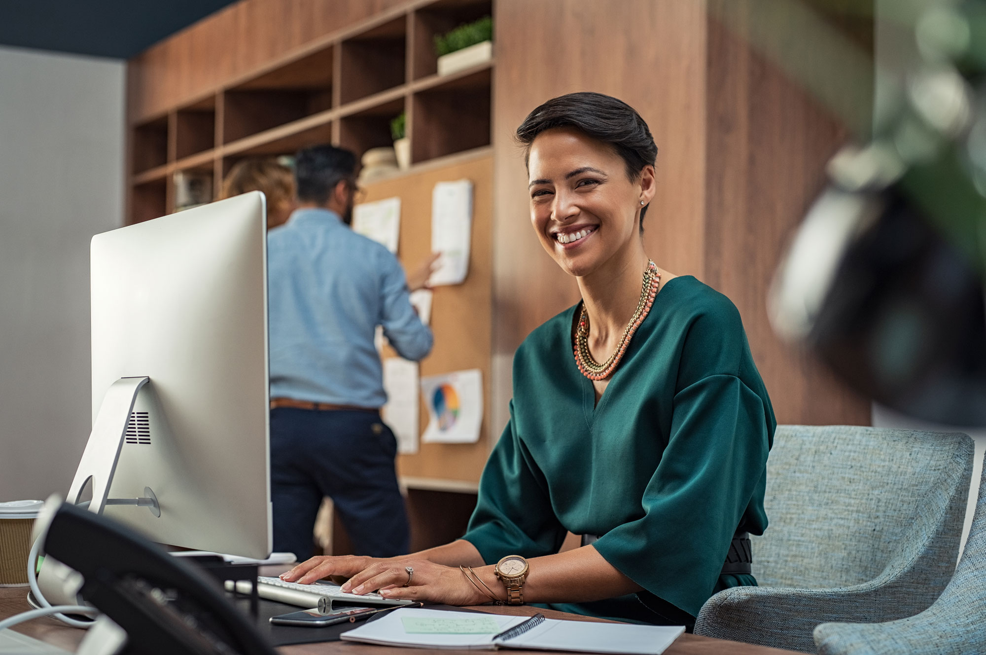 Femme d'affaires souriante travaillant sur un ordinateur au bureau avec deux collègues discutant devant un tableau au fond.