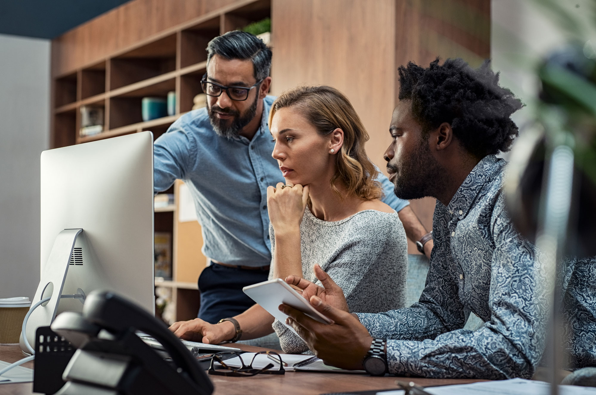 Trois collègues concentrés collaborant devant un ordinateur dans un bureau moderne avec étagères en arrière-plan.