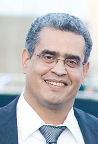 Smiling man with short curly salt-and-pepper hair wearing glasses, a white shirt, a patterned tie, and a dark jacket.