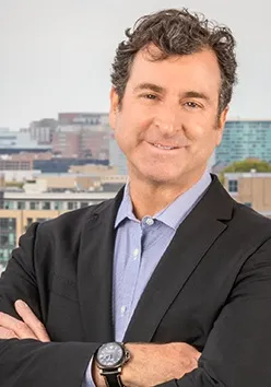 Man with curly dark hair wearing a black blazer and blue shirt, smiling with arms crossed against a cityscape background.