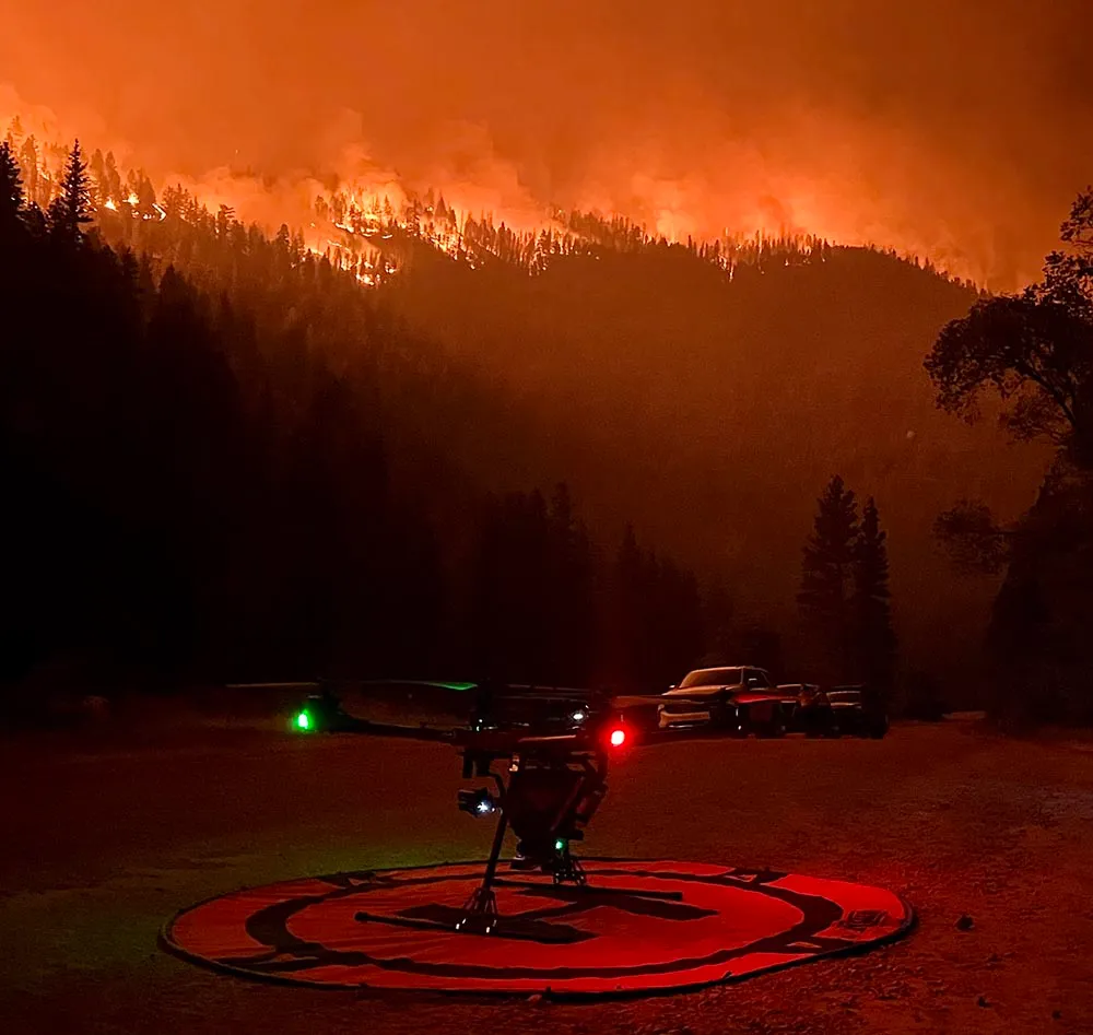 Alaris Pro | Drone with illuminated lights on a landing pad at night in front of a forest fire burning on a hillside.