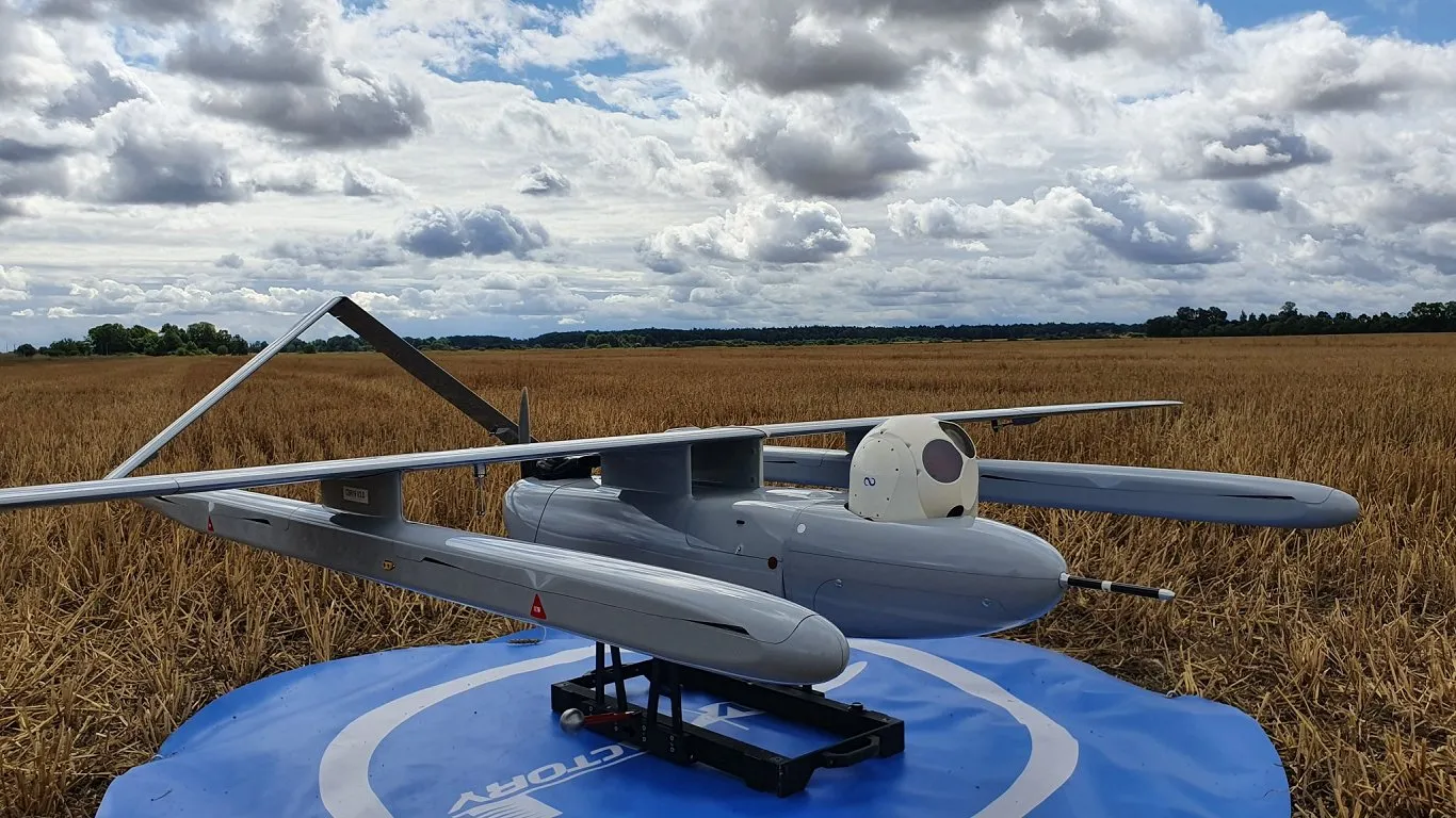 Grey unmanned aerial vehicle drone on a blue circular pad in a field under cloudy sky.