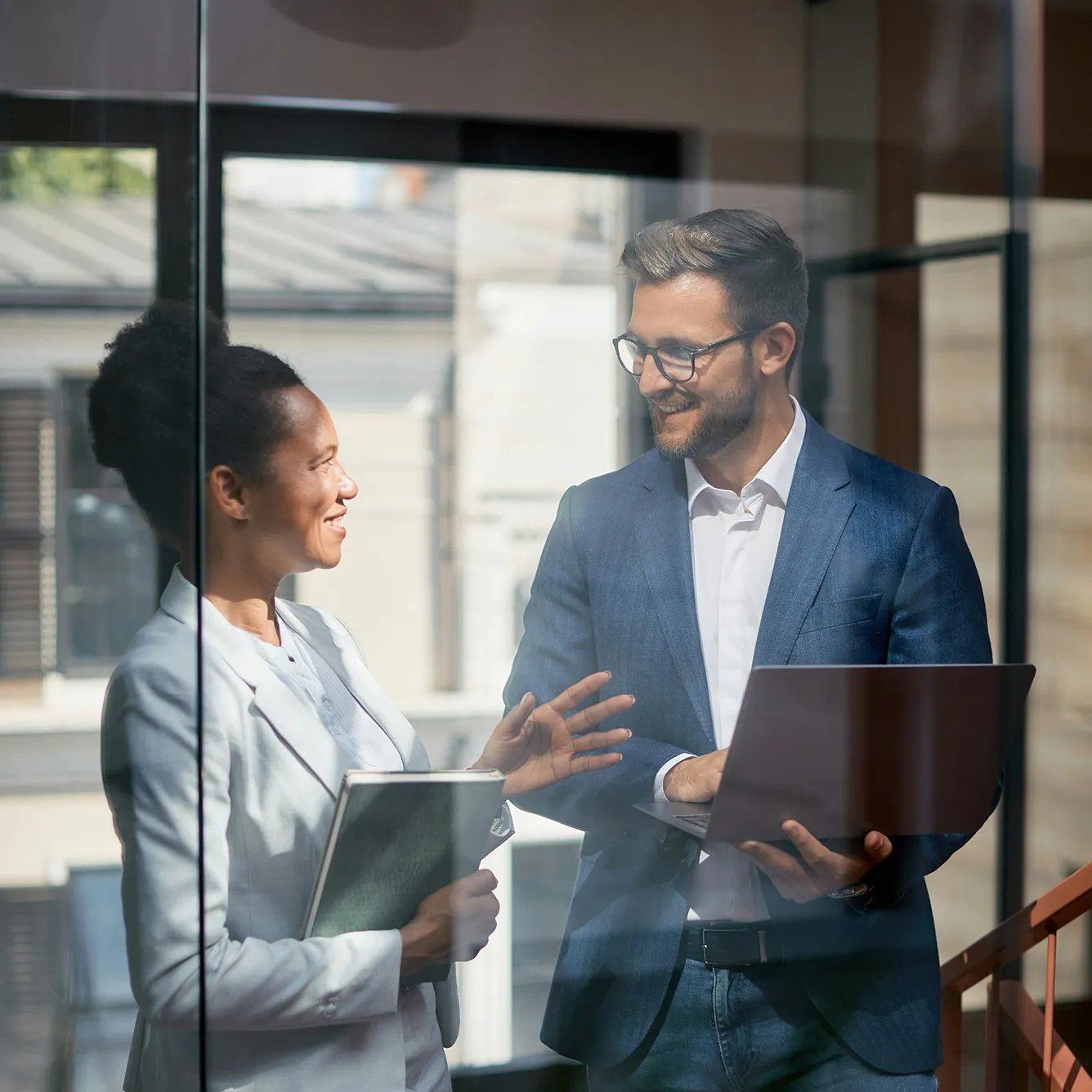 Smiling businesswoman and businessman having a discussion in an office with the man holding a laptop and the woman holding a folder.